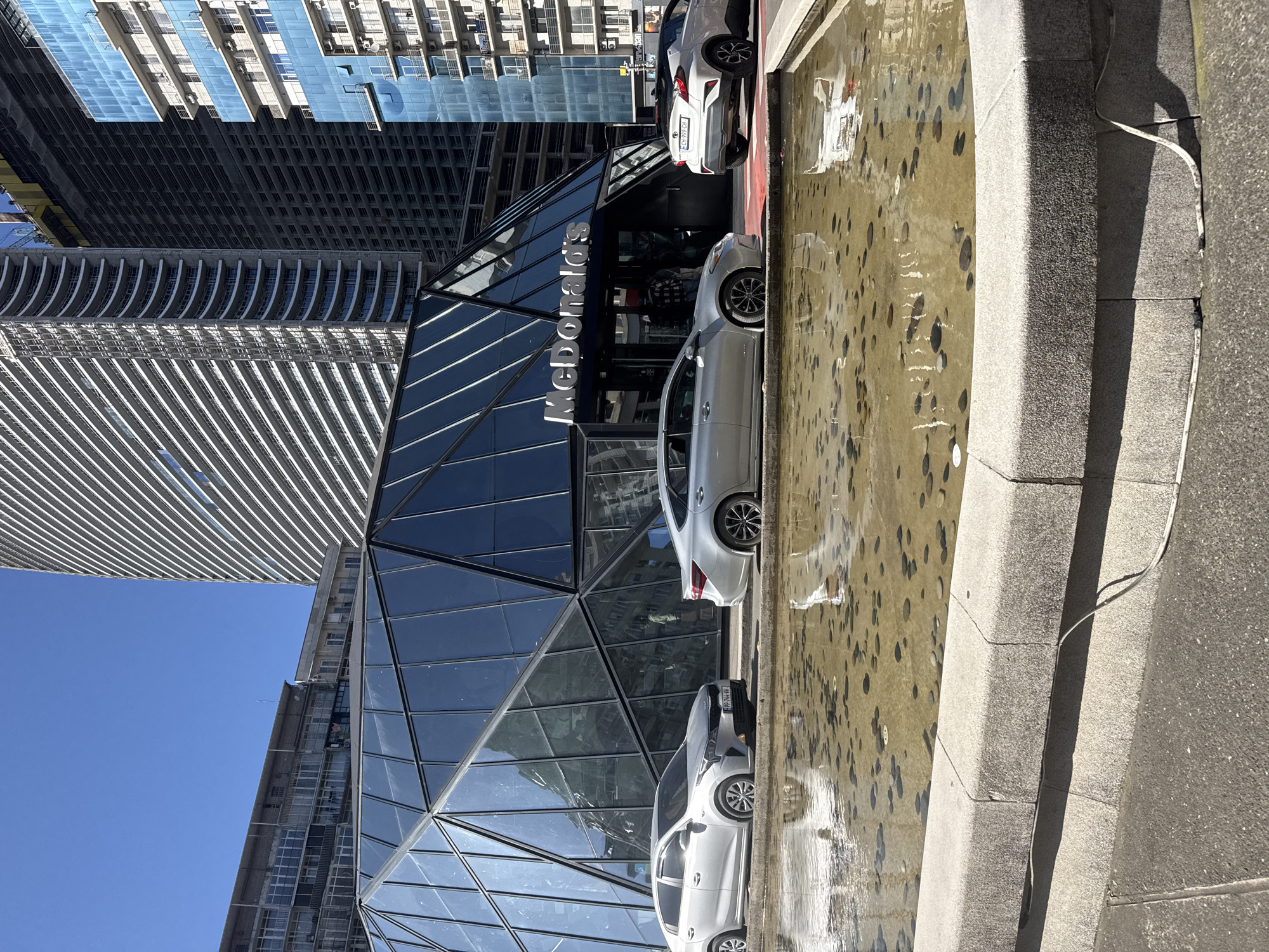 An outdoor, eye-level shot, rotated 90 degrees counter-clockwise, depicts a modern urban scene under a bright, clear blue sky. In the foreground, a rectangular fountain with shallow, murky water contains numerous coins and pebbles, suggesting it's a wishing well. A concrete ledge borders the fountain, with a white cable lying on it.
Adjacent to the fountain, several light-colored cars are parked, including a silver sedan and a white Toyota C-HR with the partial license plate "PPF 344" visible. These vehicles are positioned in front of a striking McDonald's restaurant housed within a multi-story building featuring an angular, faceted glass facade that reflects the sky and surrounding structures. The "McDonald's" logo is prominently displayed in silver lettering on the building's exterior.
In the background, a cluster of tall, contemporary high-rise buildings dominates the skyline. These structures exhibit diverse architectural styles, including one with a distinctive wavy balcony design, another with a patterned glass exterior, and others with rows of windows and visible air conditioning units. Strong shadows cast by the buildings and the parked cars indicate a sunny day. The overall scene conveys a bustling, modern urban environment with a mix of commercial and residential architecture.