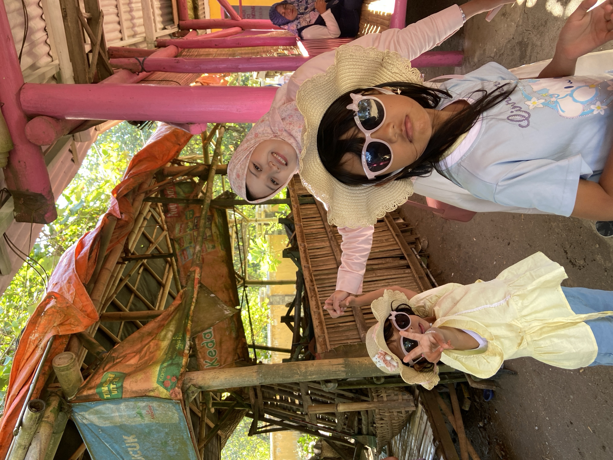 The image shows three girls posing for a photo at an outdoor location in Indonesia. The environment features a bamboo structure with tarps and various signage.
* One girl wears a pink hijab and a light pink shirt.
* Another girl wears a light blue shirt and a straw hat with sunglasses.
* A third girl wears a yellow top, blue pants, a straw hat with sunglasses, and makes a peace sign with her fingers.
There is a person in the background wearing a blue hijab. The location appears to be outdoors with trees and other structures nearby. The building is painted bright pink and is in the city of Unknown, Indonesia.