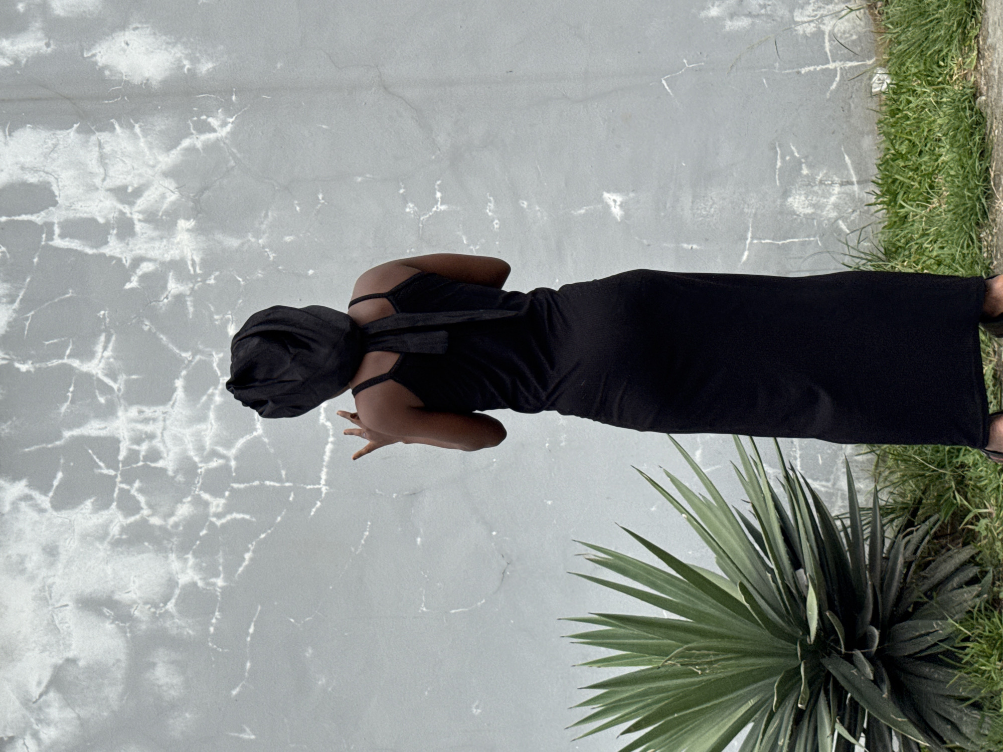 An outdoor, full-length shot features a person standing against a textured, light gray concrete wall. The person, viewed from behind and slightly to the side, wears a fitted, black sleeveless dress or jumpsuit and a black head covering. Her head is turned away from the camera, towards the wall. Her left hand is raised to chest height, fingers gently splayed.
The background wall shows prominent, light-colored crack patterns. To the right of the person, a large, dark green, spiky plant, resembling a Yucca, occupies the lower foreground. Above the plant, along what appears to be the top edge of a curb or ground, is a strip of vibrant green grass.
The photograph is taken in natural daylight. No discernible activity beyond posing is visible. The setting, with its weathered wall and tropical-looking plant, is consistent with an outdoor scene in Igboefon, Nigeria. No text is visible in the image.