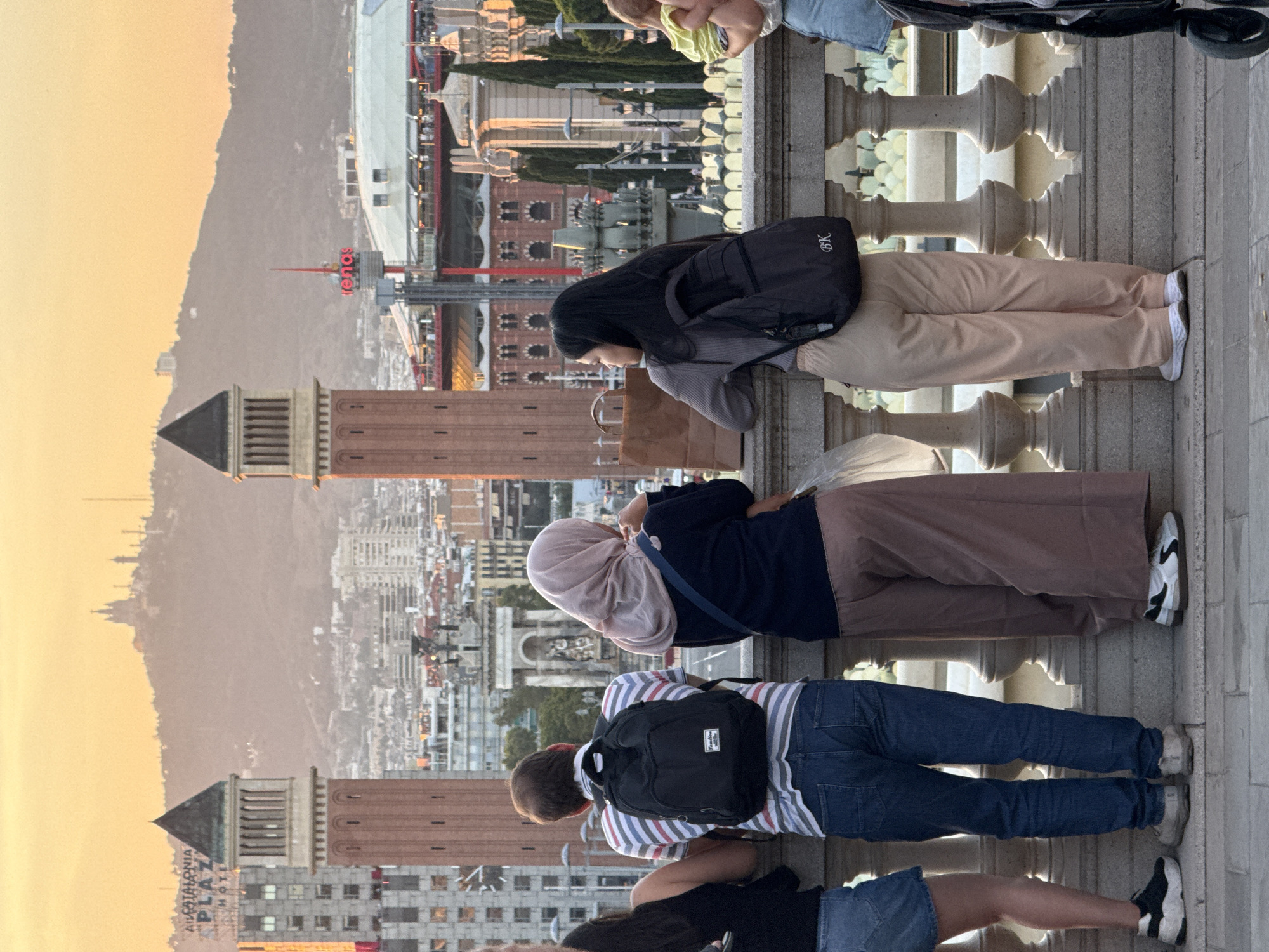 The image shows a group of people standing at a railing, looking out over a cityscape in Barcelona, Spain, at sunset. In the foreground, from left to right, are a woman with dark hair wearing a black top and a denim skirt, a man with brown hair wearing a striped t-shirt and jeans, a woman wearing a pink hijab and a dark top with loose brown pants, and a woman with long black hair wearing a grey top and light-colored pants. The man is carrying a black backpack, and the woman with black hair is also carrying a black backpack. Behind the people is a wide, ornate railing with decorative pillars. The background features a panoramic view of Barcelona. Prominent in the mid-ground are two tall, red brick towers with dark pyramidal roofs, possibly part of the Venetian Towers (Torres Venecianes) near Plaça d'Espanya. Beyond them, the city stretches out with numerous buildings, including some with distinctive architectural styles. In the far distance, a large hill with a building or monument at its summit is visible against an orange sky, characteristic of sunset. A large, modern building with a white roof and red accents is also visible in the mid-ground, possibly a venue like "Las Arenas de Barcelona". The overall scene captures a moment of observation and appreciation of the city's skyline.