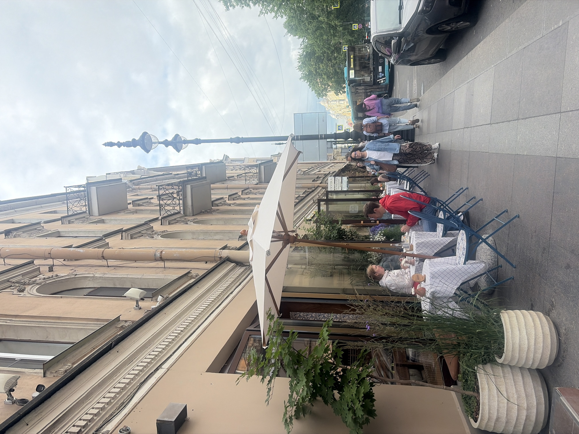 The image depicts an outdoor seating area of a cafe on a cloudy day in Saint Petersburg, Russia. Several people are visible, some walking on the sidewalk and others seated at tables covered with checkered tablecloths. One person in a red jacket is leaning over a table, while another person in a white jacket is seated. A large white umbrella shades the seating area. The background shows a building with ornate architecture, a street with a bus and cars, and trees lining the sidewalk. The overall atmosphere appears to be relaxed and casual, with a hint of urban life. The time of day is likely daytime, given the natural light. There is no visible text in the image.