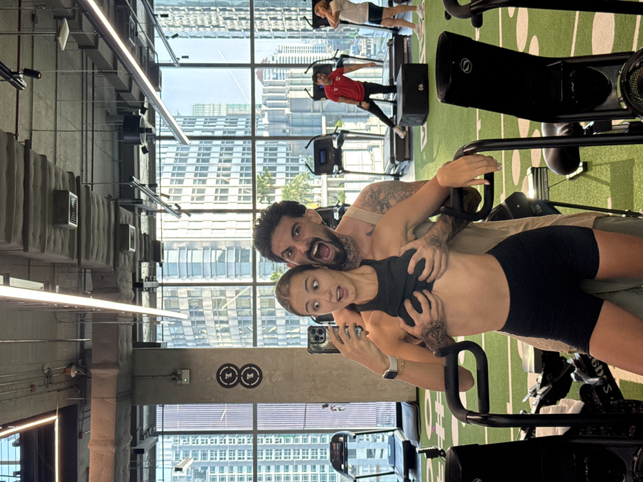 An interior photograph depicts a modern gym located in Bangkok, Thailand, with a panoramic view of a city skyline through large windows.
In the immediate foreground, a Caucasian female, approximately 20-30 years old with light brown hair, is centered. She is wearing a black sports bra and black shorts, holding a silver-colored smartphone in her left hand, appearing to take a selfie. Her expression is wide-eyed with an open mouth, conveying surprise or playful alarm. Her right hand rests on the handlebar of a black exercise machine. Directly behind her, a male, also approximately 20-30 years old with dark hair, a beard, and visible tattoos on his neck and arms, embraces her. He wears a light-colored sleeveless tank top. His hands are positioned underneath the woman's sports bra, appearing to lift it slightly. He displays an exaggerated open-mouthed expression, resembling a scream or roar.
The gym floor features a green surface with white markings. Various gym equipment is visible, including multiple black treadmills, a black plyo box, and other exercise machines. To the right of the central figures, another male in a red t-shirt and black shorts is observed performing an exercise, with one leg extended onto the plyo box. Further to the right, a second female in a light-colored t-shirt and dark shorts stands. A concrete pillar on the left wall features two circular, stylized '8'-shaped logos. The ceiling is exposed concrete, fitted with linear LED light fixtures and visible ventilation ducts. Beyond the expansive windows, numerous high-rise buildings under a clear sky form a typical dense urban landscape.