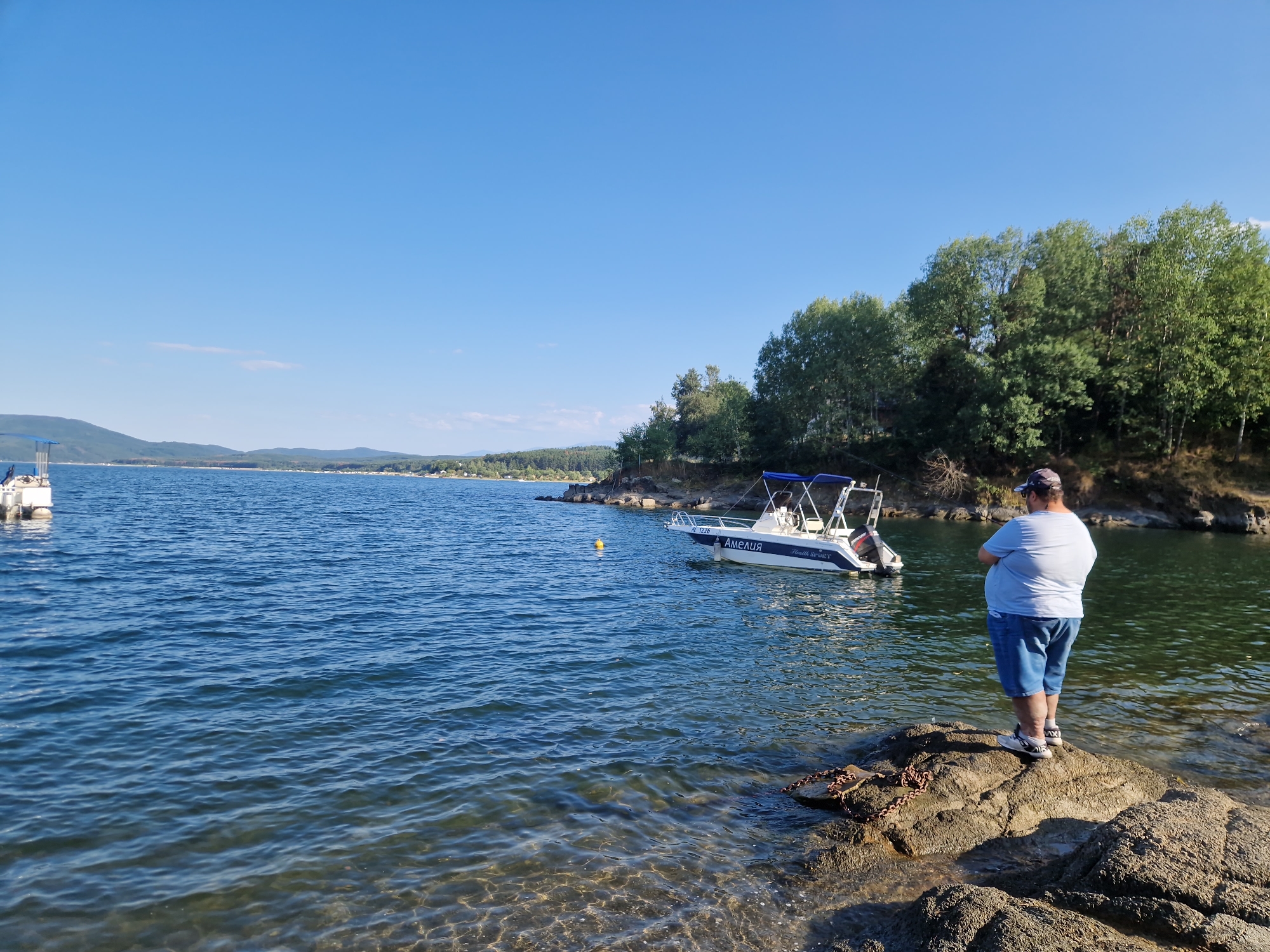 In the city of Dolni Pasarel, Bulgaria, a man stands on a rocky shore overlooking a calm body of water. The man, wearing a light blue t-shirt and denim shorts, is facing away from the camera, looking out at the lake. He has a cap on his head and is wearing athletic shoes. Nearby, a white motorboat with a blue canopy is docked. The water is clear and reflects the bright blue sky, with a few small waves rippling its surface. In the distance, a line of green trees borders the far side of the lake, with rolling hills visible behind them. The sun is shining brightly, casting shadows on the rocks and water, indicating it is likely daytime.