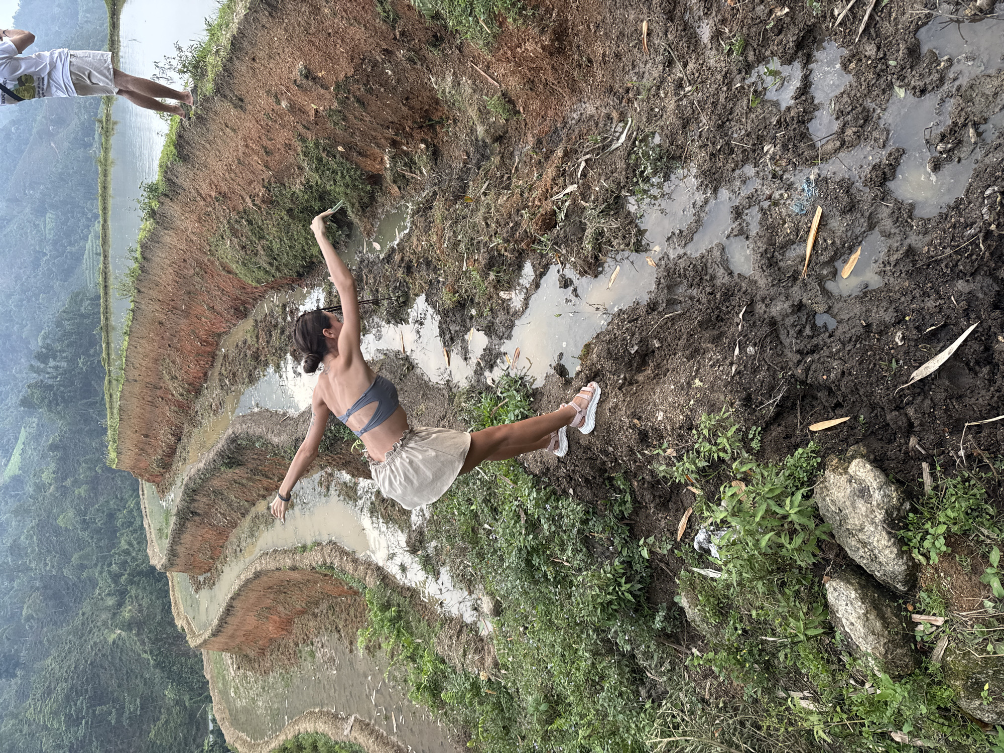 The image depicts a vibrant, mountainous landscape in Nậm Dịch Commune, Vietnam, featuring terraced rice paddies. The scene is captured during daytime under an overcast or hazy sky, suggesting a humid climate.
In the foreground, a woman is balancing on a muddy, unpaved edge overlooking the terraces. She wears a gray bralette or swim top, a light-colored, ruffled skirt, and white platform sandals. Her arms are outstretched, and one leg is lifted behind her, suggesting a pose of joy, freedom, or dance. She holds a mobile phone in her right hand, possibly taking a selfie or recording.
Behind her, partially visible to the left of the frame, stands a man. He is wearing a white t-shirt with indistinct text on it and light-colored shorts, also with sandals. He appears to be observing the scene or the woman.
The immediate surroundings feature dark, wet soil with puddles and sparse green vegetation, indicating a muddy path or field edge. Beyond this, the landscape descends into multiple layers of terraced rice paddies, filled with water, reflecting the sky. These terraces curve along the contours of the hills. In the far background, lush green mountains covered in dense vegetation rise under the soft, diffused light.
The photograph captures a moment of engagement with the scenic agricultural environment, characterized by the iconic terraced rice fields.