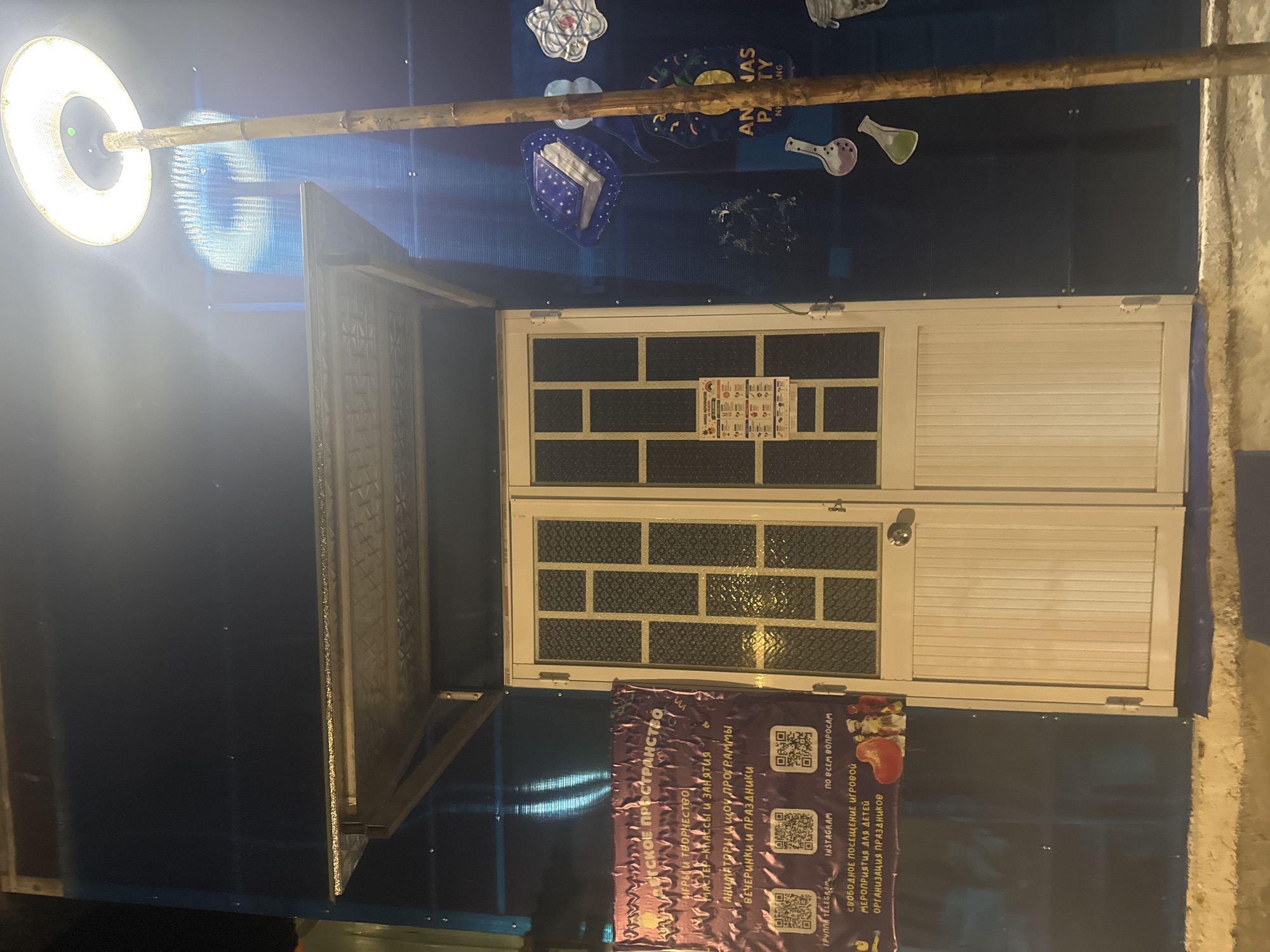 The image captures an outdoor scene at night in Bac Nha Trang Ward, Vietnam. A building entrance is visible, featuring double white doors with a grid pattern on the upper half. Above the doors, a metal awning provides shelter. To the left of the entrance, a purple banner displays text in Russian, advertising a children's space with games, art, masterclasses, and party organization. QR codes and social media handles are also present on the banner. To the right of the entrance, the wall is adorned with blue decorations, including what appears to be a book with a starry cover and other whimsical shapes. A tall, thin bamboo pole stands vertically in front of this wall, supporting a round, illuminated outdoor heater overhead. The lighting creates a stark contrast between the illuminated objects and the darker background. The ground in front of the building appears to be a rough concrete or stone surface.
