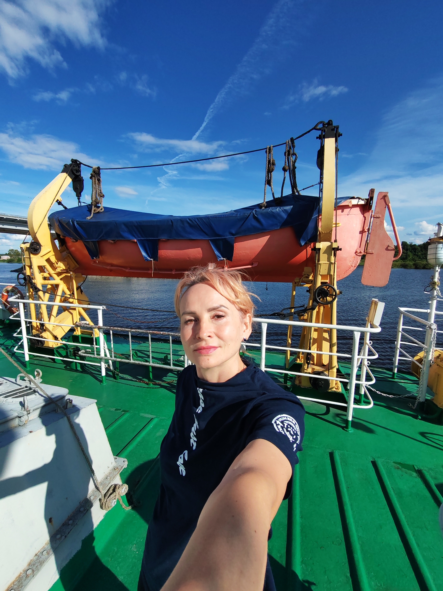 A woman with short, light-colored hair takes a selfie on the deck of a vessel. She is wearing a dark blue t-shirt with a circular emblem on the left sleeve. She is smiling slightly and looking at the camera.
Behind her, a bright orange and red lifeboat is suspended from a yellow davit. The lifeboat is covered with a blue tarp. The deck of the vessel is painted green. A white railing with a chain runs along the edge of the deck. In the background, there is a body of water, a bridge, and the shoreline with trees.
The sky is blue with scattered white clouds. The sun is shining, suggesting it is daytime. The scene appears to be in Sheksna, Russia, based on the provided location context. The overall mood seems calm and pleasant. There is no visible text in the image, other than what is on the emblem on the woman's t-shirt, which is not legible.