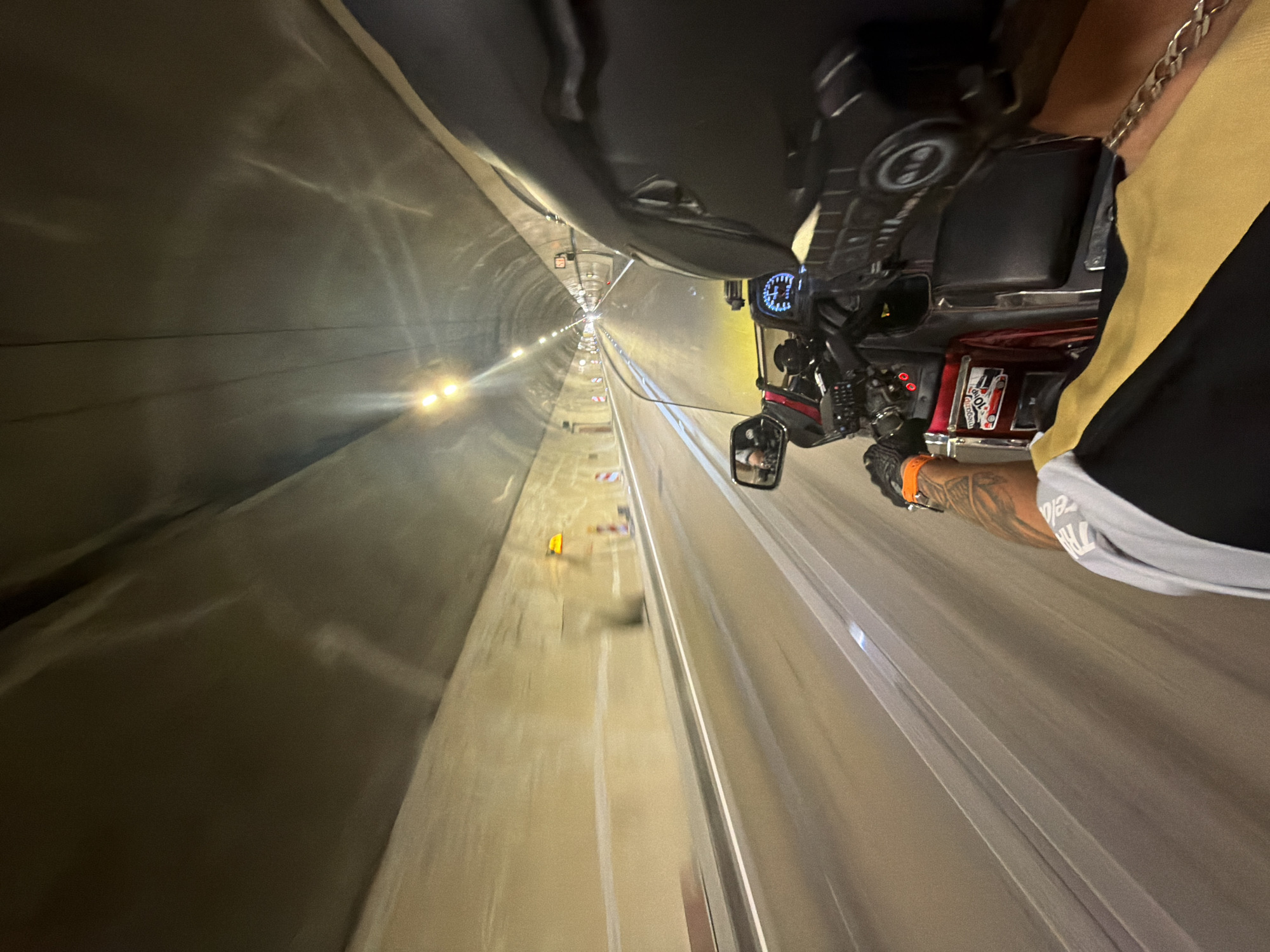 The image is a first-person perspective shot from a motorcycle traveling through a tunnel. The tunnel walls are made of a light-colored material and are illuminated by overhead lights. There are white lines marking lanes on the road. The rider's arm is visible in the foreground, wearing a black glove with an orange band and a tattoo on the forearm. The motorcycle's dashboard is also visible, with a speedometer and other gauges. The scene appears to be in motion, with a slight blur indicating the motorcycle's movement. The location is stated to be in the city of Đurmani, Montenegro, though this specific detail cannot be verified from the visual information alone.