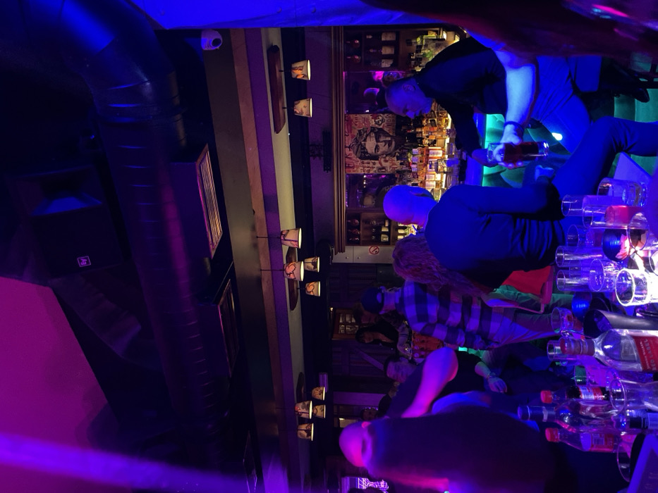 The image, taken in Drapetsona, Greece, captures a vibrant indoor bar or nightclub scene at night, bathed in striking purple and blue neon lighting. The setting features multiple individuals gathered around a bustling bar area.
In the background, two people, likely bartenders, are positioned behind a well-stocked bar. One individual, with a light or bald head, stands upright, while another leans forward, holding a bottle and appearing to pour or serve drinks. Behind them, the bar shelves are filled with an extensive collection of liquor bottles and various types of glassware. A prominent piece of framed artwork or a poster featuring a stylized portrait of a man hangs on the wall behind the bar.
In the midground and foreground, several patrons are visible, though their faces are largely obscured by the low light and camera angle. One person can be seen wearing a plaid shirt, and others have long hair. The foreground of the image showcases a bar top laden with numerous stacked and scattered glasses, some appearing empty, alongside various bottles. Notably, a label on one of these bottles clearly displays the text "WALKER," suggesting a Johnnie Walker whisky bottle.
The establishment exhibits an industrial aesthetic with exposed dark ductwork running along the ceiling. A white security camera is mounted on the ceiling in the upper left corner. Along a wall, several decorative, wall-mounted lamps with patterned shades emit a softer, warmer glow that contrasts with the predominant neon. A dark speaker, bearing the "EV" (Electro-Voice) logo, is also visible, indicating a sound system. The overall atmosphere conveys a lively and social gathering typical of a nightlife venue.