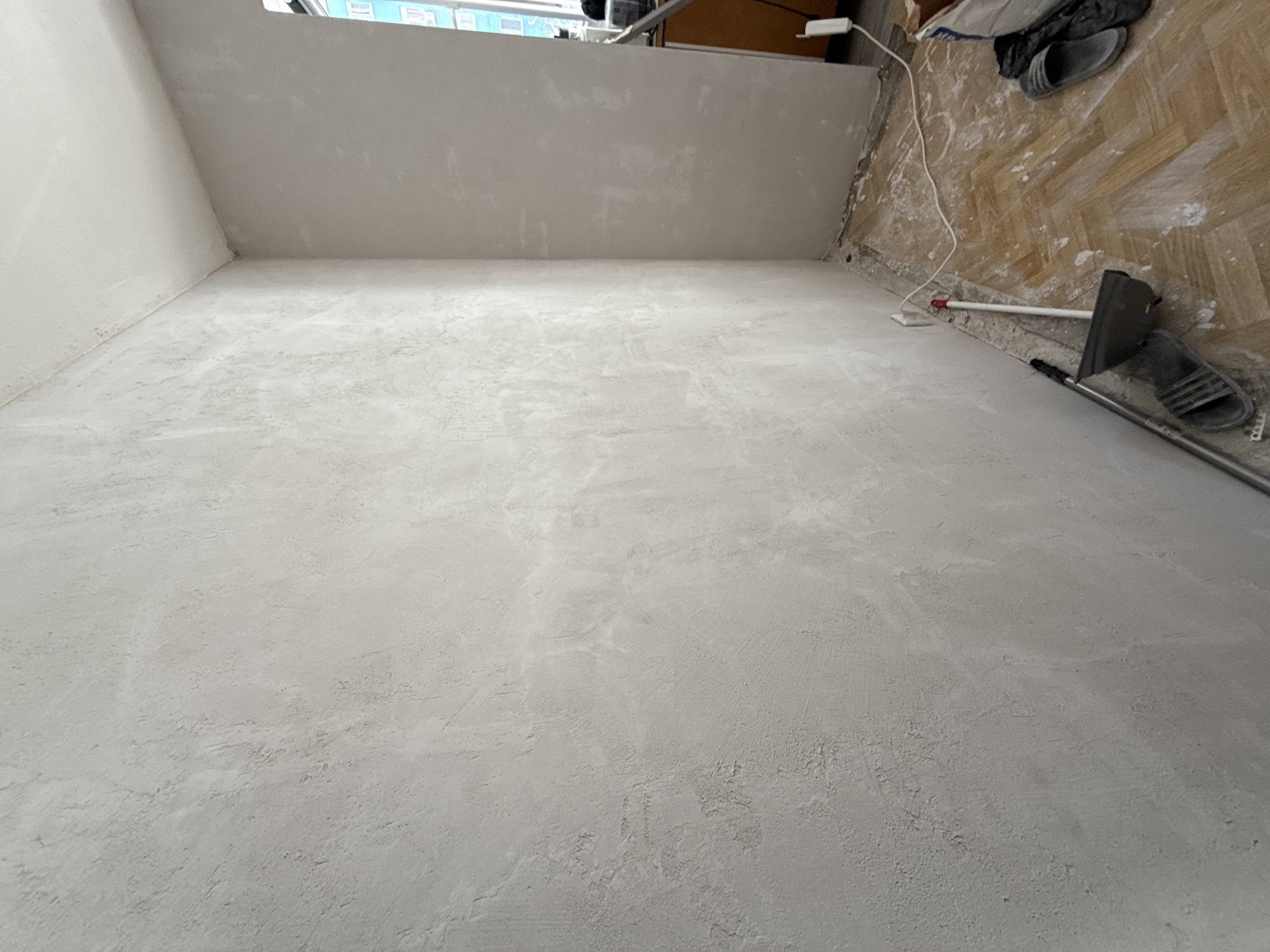 The scene depicts an interior space undergoing renovation, likely in Stara Zagora, Bulgaria (city inferred based on provided location). The primary focus is on a room with walls and a floor covered in a freshly applied, rough-textured grey coating (possibly plaster or a similar material). The room has a corner where the walls meet, and the top of one wall extends to a window or opening, suggesting natural light is present.
To the right of the image, the floor is visible showing a herringbone pattern hardwood floor partially covered with white powder, potentially from the renovation process. Near the wall, there are objects including a power cord, a pair of grey sandals, a bag, and what appears to be a plastering or skimming blade attached to a long handle resting on the new grey surface. The overall impression is one of a work in progress, with the space in a state of transition from its original state to a renovated condition. There are no people depicted.