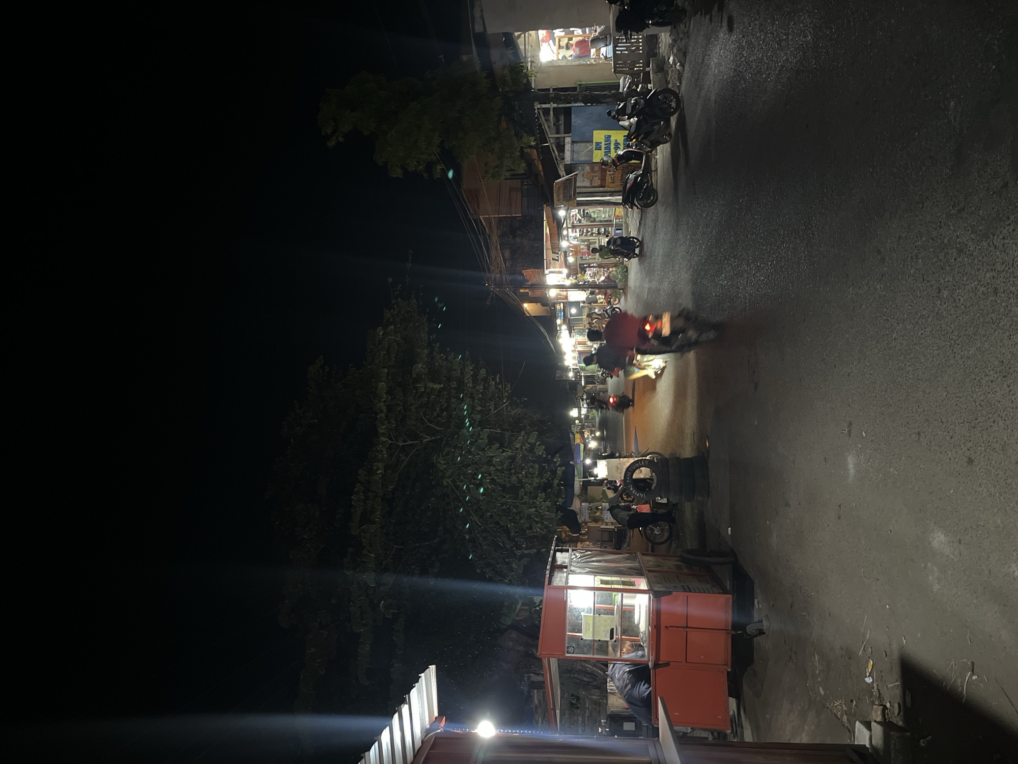 The image depicts a street scene at night in Pecangaan, Indonesia, characterized by low-light conditions and artificial illumination from shops and streetlights. A red food cart is prominent on the left side of the street, with a person visible inside it. Several motorcycles are present, with some in motion and others parked along the roadside. Figures of people can be seen on and around the motorcycles, as well as in front of the illuminated shops lining the street. The street appears paved, and vegetation, including trees, is visible in the background. The overall atmosphere suggests a commercial area with activity continuing into the evening.