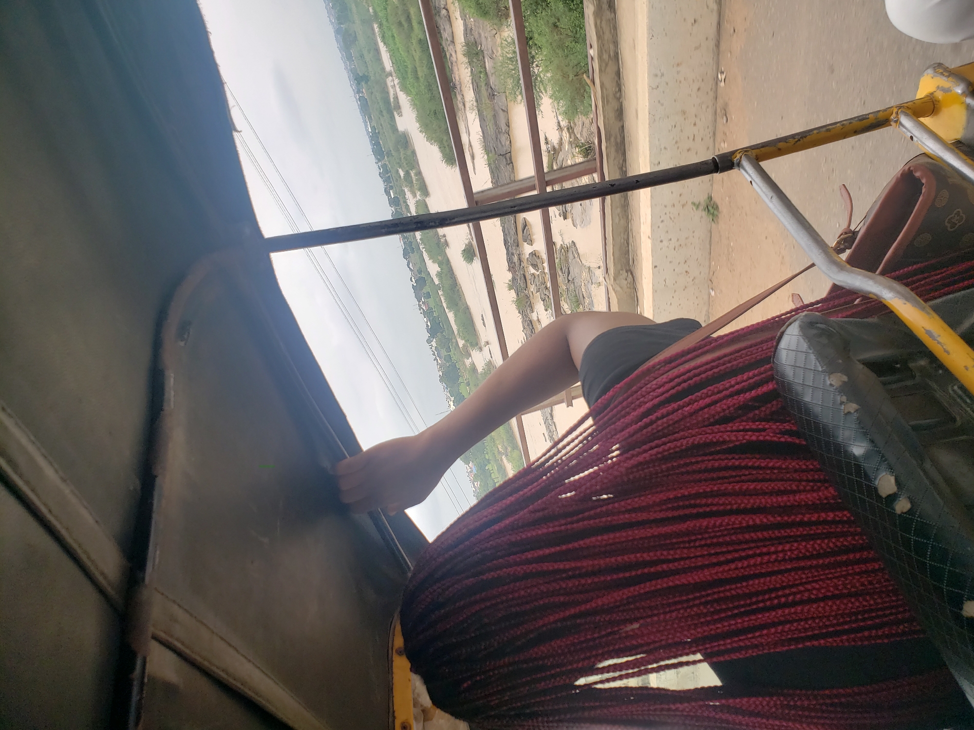 A rear view of a person with long, red braided hair sits in the passenger seat of a yellow auto rickshaw, facing the right. Their right hand is visible, gripping the roof of the auto rickshaw. Outside the vehicle, a wide, muddy river flows between rocky banks and is lined with green vegetation. In the distance, buildings and power lines are visible, suggesting an urban or semi-urban environment. The sky is overcast, and the overall lighting is diffused.