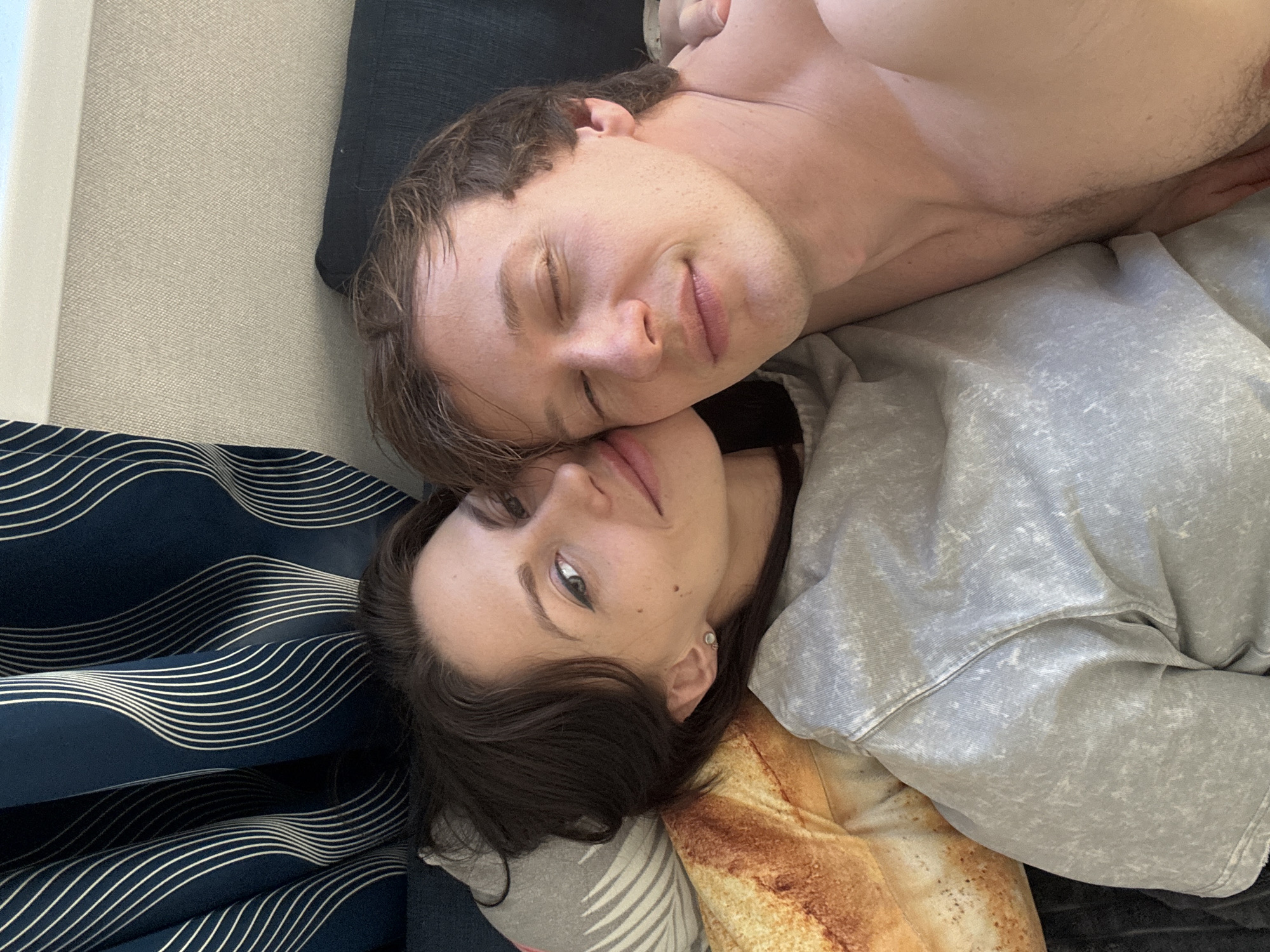 Two individuals are lying close together. One appears to be a man with his eyes closed, shirtless, with brown hair. The other appears to be a woman, awake, with dark hair. They are in a bed with patterned bedding and pillows. The location cannot be determined to be in Murino, Russia.