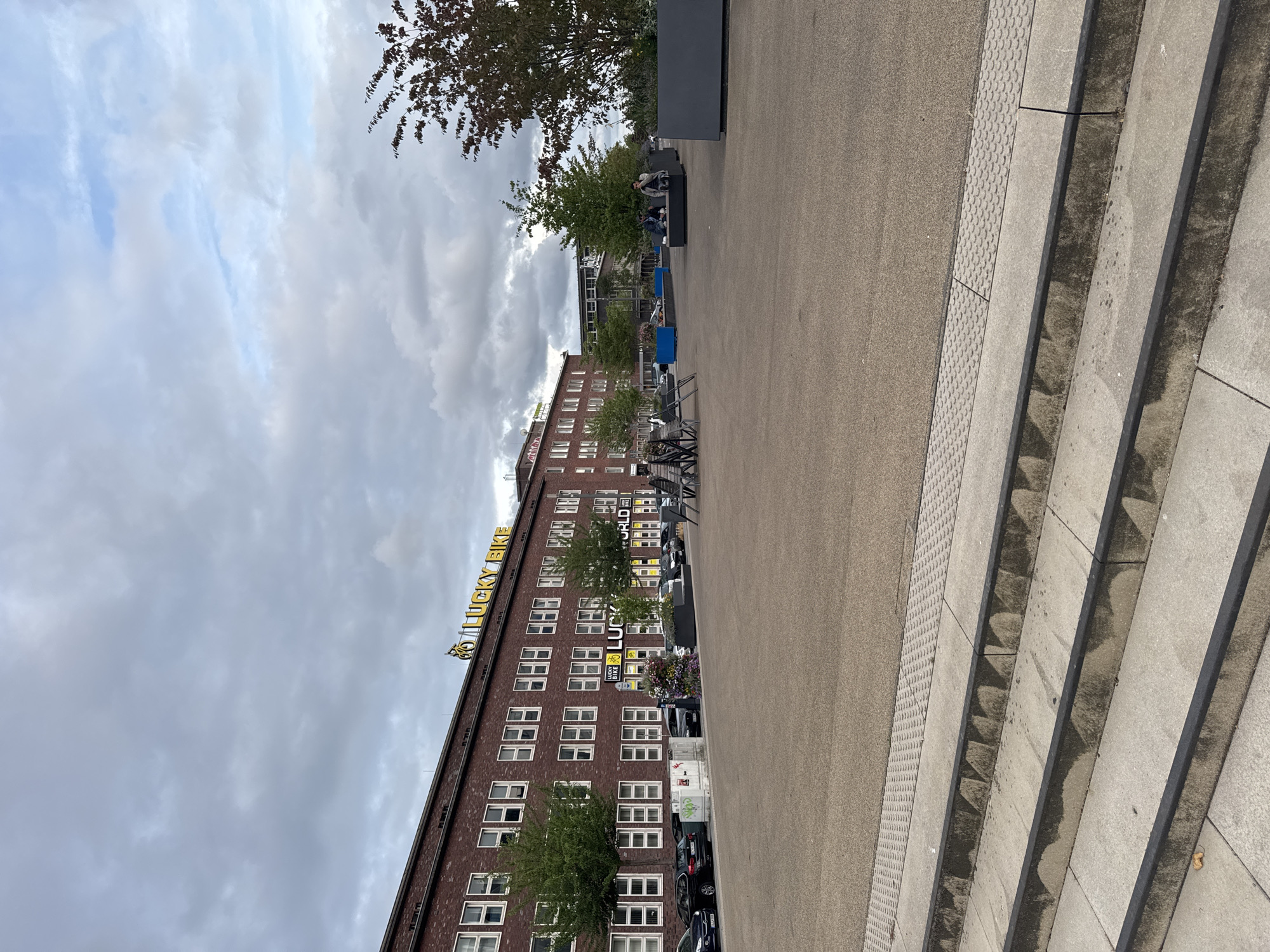 This image depicts a street scene in Duisburg, Germany, specifically at the Lucky Bike store located at the former site of the Thyssen Krupp Steelworks. In the foreground, a set of concrete stairs leads up to a wide, open paved area. The paved area is populated by several dark grey planter boxes and some modern outdoor seating areas with benches and chairs. Two individuals are seated on a bench in the middle-ground, appearing to be engaged in conversation. Behind them, the brick facade of a large building is visible, adorned with a prominent yellow "LUCKY BIKE" sign and a bicycle graphic. Several cars are parked along the street in front of the building. The sky above is overcast with scattered clouds, suggesting a cloudy day. The overall environment appears to be a contemporary urban space with a mix of commercial and public areas.