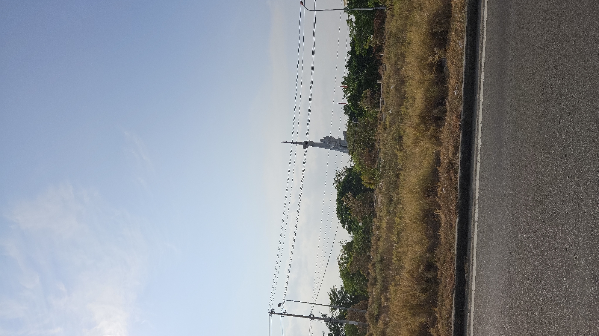The image depicts a landscape in Cam Ranh, Vietnam. A road with a gray asphalt surface runs along the right edge of the frame. Adjacent to the road is a strip of dry, golden grass and some green vegetation. In the distance, a concrete monument with a tall, pointed spire stands out against the blue sky. Power lines stretch across the sky. A streetlight is visible in the lower left corner. The sky is predominantly blue with some faint wisps of clouds.