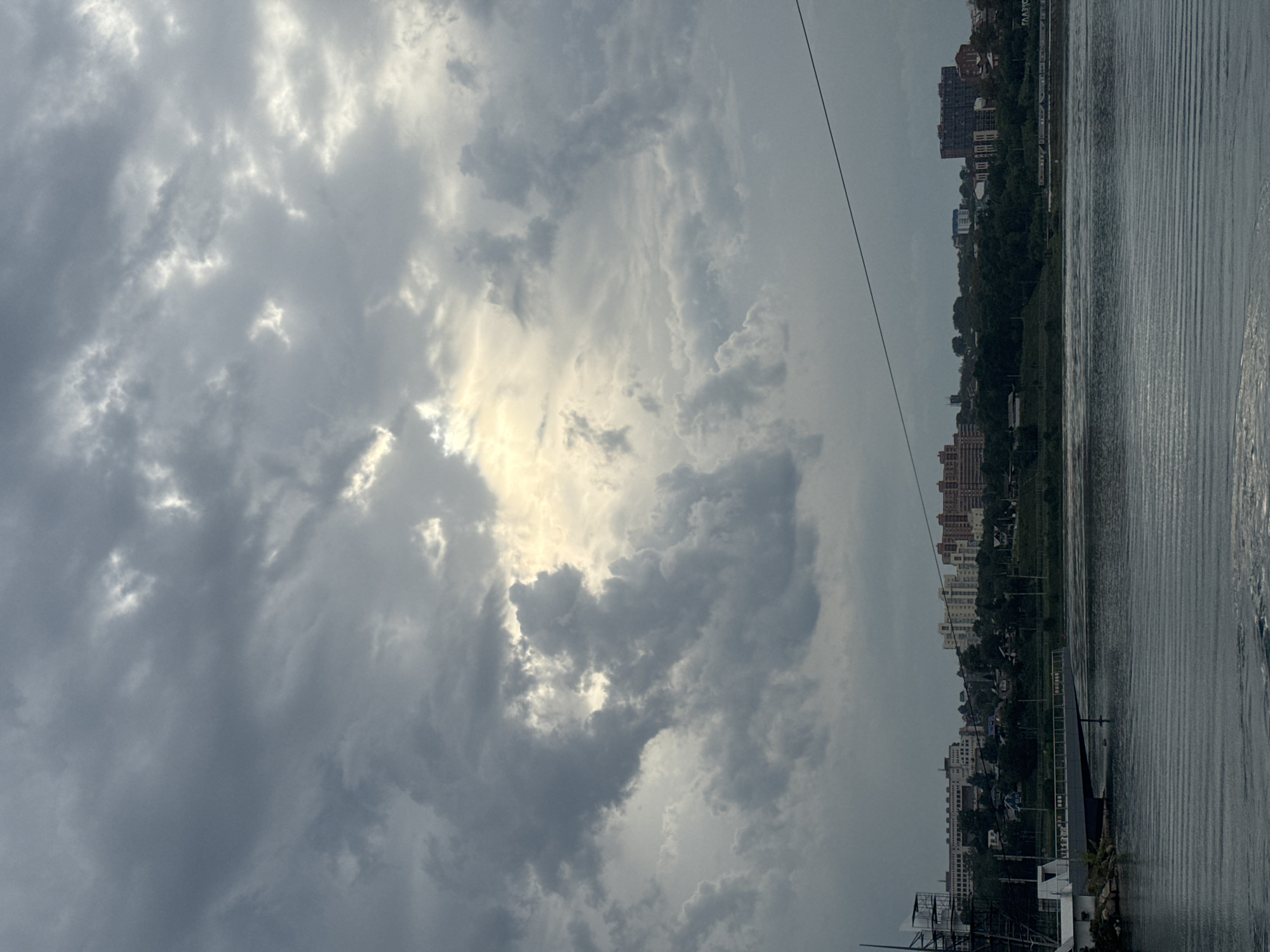 The image captures a cloudy sky over the Angara River in Irkutsk, Russia. On the riverbank, several buildings, including multi-story apartment complexes and smaller houses, are visible, nestled amongst lush green trees. A prominent structure on the left appears to be a part of a large boat or ship, possibly a museum or a historical vessel, with scaffolding around it. A bridge extends into the water from the bank. The water itself is rippled, reflecting the overcast sky. A thin wire runs across the upper portion of the image.