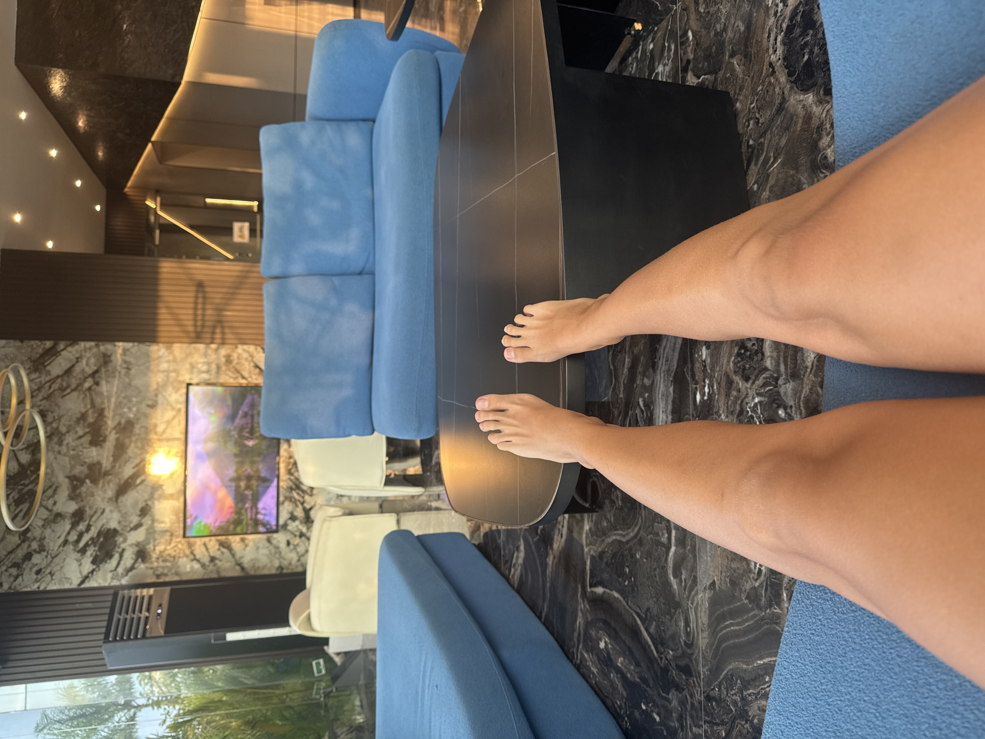 The image displays a modern, upscale lounge area from a first-person perspective, focusing on a person's legs and bare feet in the foreground. The individual is relaxed, with their legs extended over a dark, sleek coffee table that occupies the central mid-ground.
The setting appears to be a contemporary living room, hotel lobby, or lounge. Two plush, light blue sofas are partially visible, one on either side of the frame, suggesting the viewer is seated on one. Further back, two cream-colored armchairs are arranged, contributing to the seating ensemble. The flooring throughout the space is a luxurious dark marble or stone with prominent white veining.
On the wall in the background, a flat-screen television displays a vibrant, abstract image with purple, pink, and green tones. Below the TV, a long, black soundbar or air conditioning unit is integrated into the wall design. To the left of the television, an abstract metallic art piece, composed of intertwined rings, adds a decorative touch. The walls themselves feature a mix of dark patterned panels and lighter sections with recessed spotlights, creating a sophisticated ambiance. Warm lighting illuminates the space, suggesting either natural light from a window or well-placed artificial lighting, possibly indicating late afternoon or early evening. Reflections of green foliage, likely palm trees, can be seen in a large window or glass partition in the bottom left, hinting at a tropical outdoor environment. A small, unidentifiable symbol or text is faintly visible on a background panel. The overall scene suggests comfort and modern elegance, aligning with the location context of North Kuta, Indonesia.