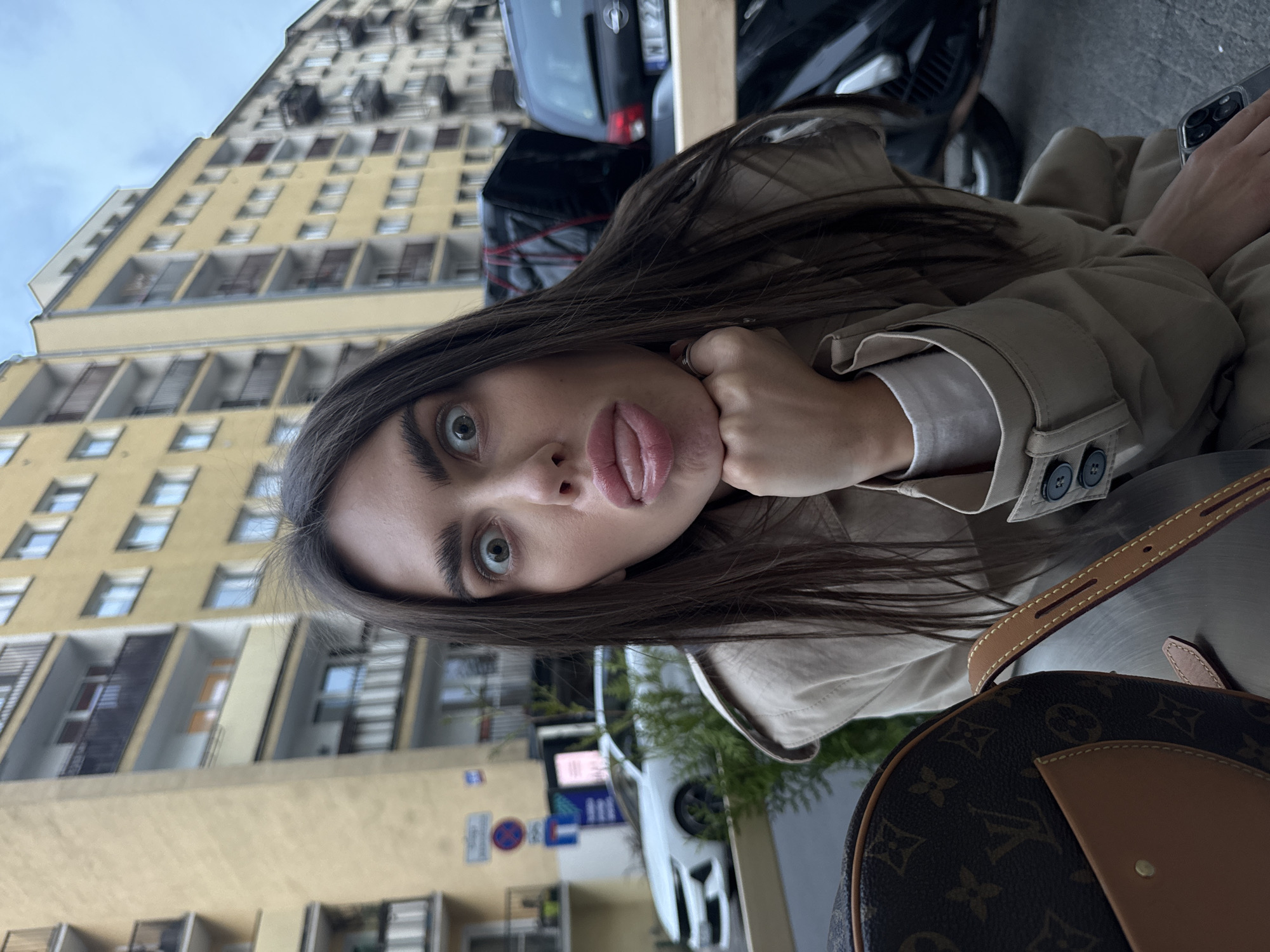 A woman with dark hair, fair skin, and green eyes is posing for a selfie. She is wearing a light-colored trench coat and resting her chin on her hand. Her expression is playful, with her tongue slightly protruding. Part of a Louis Vuitton handbag is visible in the lower right corner of the frame.
The background features a tall, yellow building with numerous windows. Cars are parked along the street, and street signs are visible. Based on the architectural style of the building and the appearance of the street signs, the location is likely in Warsaw, Poland.