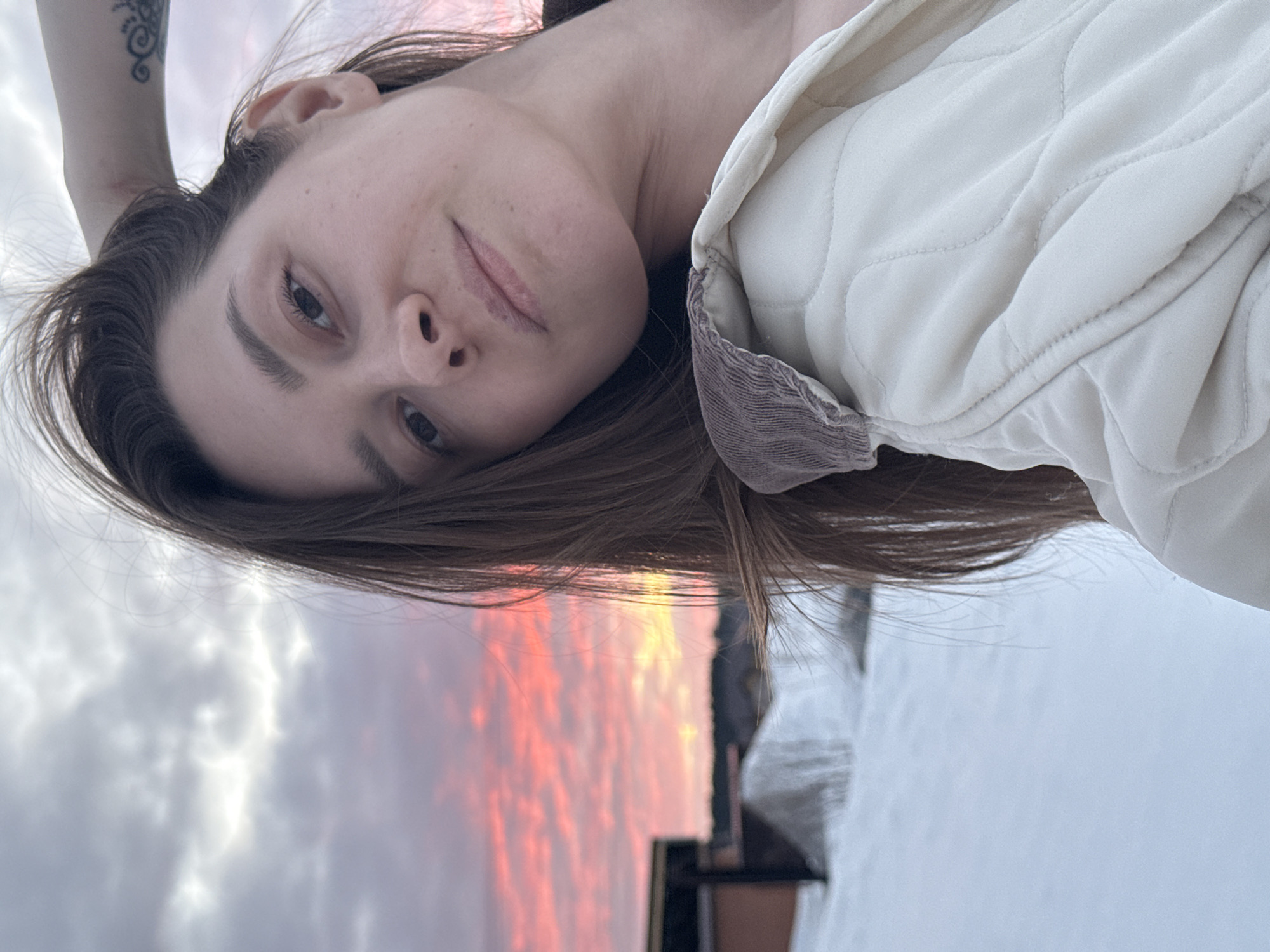 A young adult female is depicted in a close-up outdoor selfie in Chaikovsky, Russia. Her head is tilted slightly, and she looks directly at the camera with a subtle smile. She has long, dark brown hair, with strands visible across her face and shoulder. Her left hand is raised, fingers touching her hair near the top of her head, revealing a dark, coiled tattoo on her left upper arm. She wears an off-white quilted jacket with a distinct brown corduroy collar. The background features a snowy landscape under a dramatic sunset sky, exhibiting vibrant pink, orange, and red cloud formations that transition into lighter gray and blue tones overhead. In the blurred mid-ground, behind the subject, structures with dark frames and reddish-brown facades are visible amidst snowdrifts.