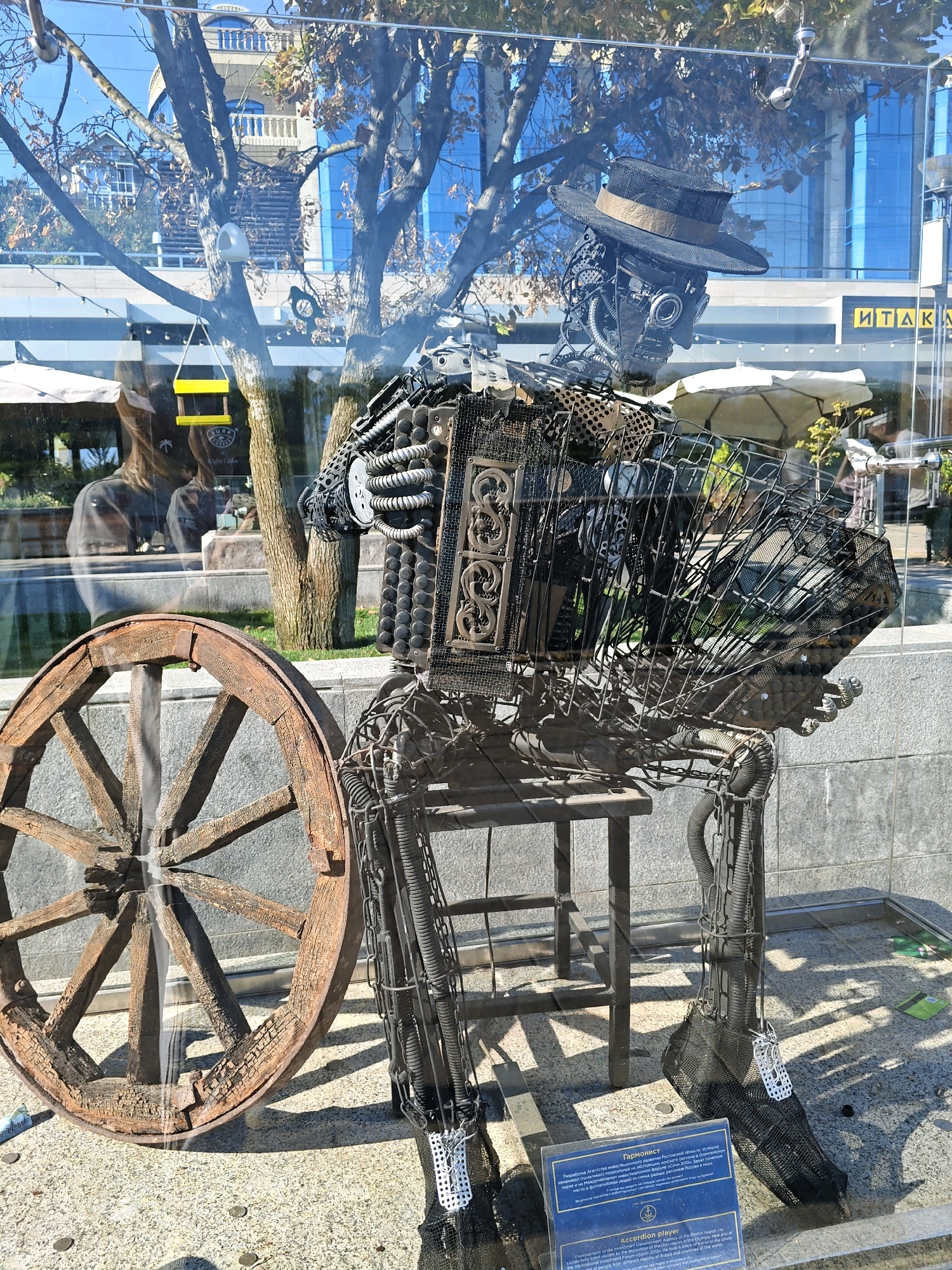 The image displays a robotic sculpture of an accordion player placed behind a glass barrier in an outdoor urban setting, likely a public space or a shop window in Rostov-on-Don, Russia. The sculpture is constructed from metal, incorporating elements such as springs, gears, and wire mesh, and is adorned with a hat. To the left of the sculpture is a large, weathered wooden wheel. In the background, trees with autumn foliage are visible, along with modern buildings featuring blue glass facades and signage. A blue informational plaque with text in both Russian and English is positioned at the base of the sculpture, identifying it as "Гармонист" or "Accordion player" and detailing its connection to the Rostov region, Russia. Sunlight casts shadows on the ground.