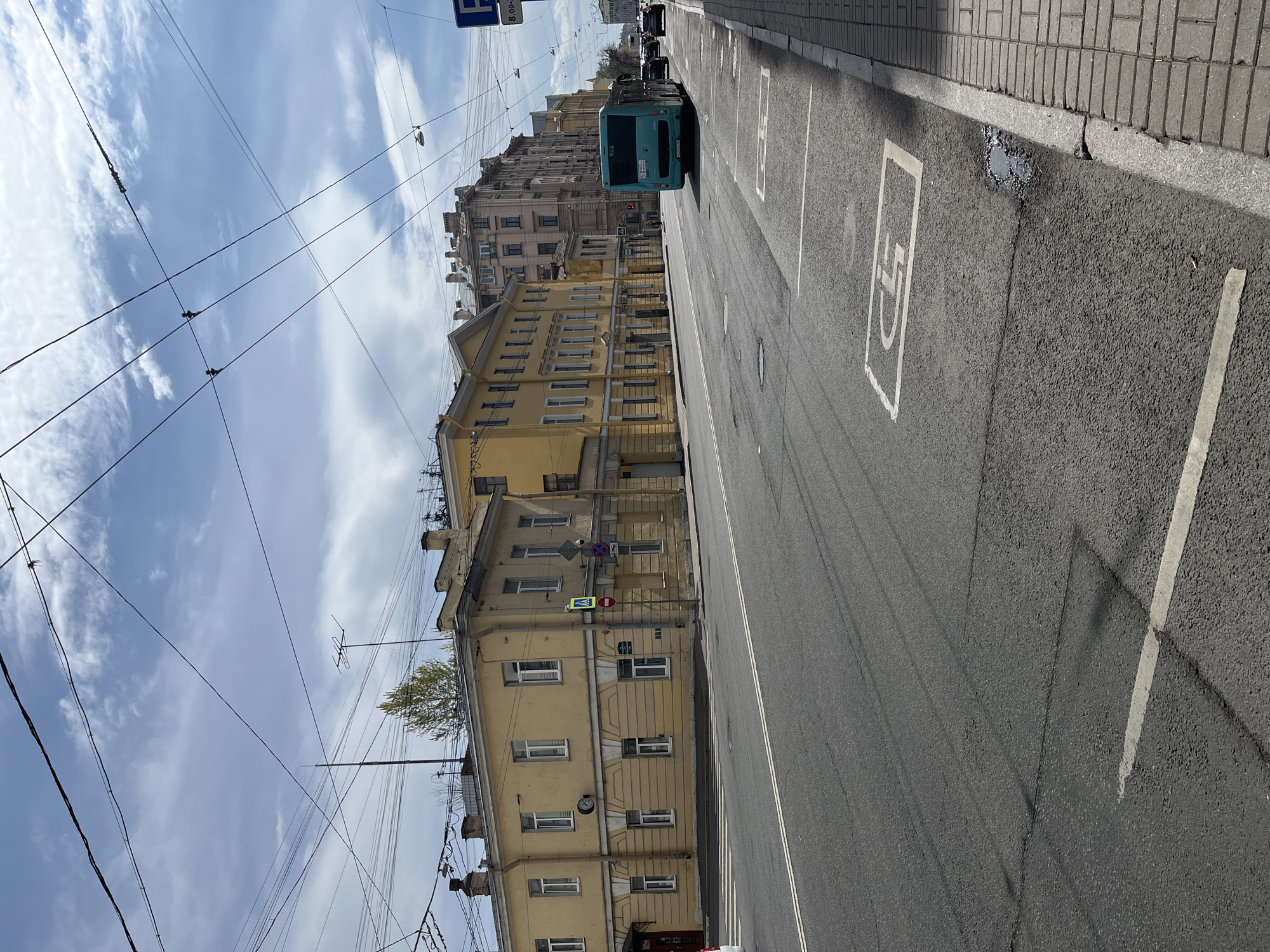 The image depicts a street in Saint Petersburg, Russia. The perspective is ground level, looking down the road with a slight tilt to the left. On the right side of the street is a narrow sidewalk paved with rectangular blocks. The road itself is asphalt and marked with parking spaces, one displaying the handicapped symbol. On the left, a yellow building with white-framed windows stands tall, its facade revealing multiple stories. Overhead, a network of utility wires crisscrosses against a cloudy sky. A blue bus is seen driving away from the camera in the distance, adding a sense of motion to the urban scene. Traffic signals are visible near the yellow building, suggesting an intersection nearby.