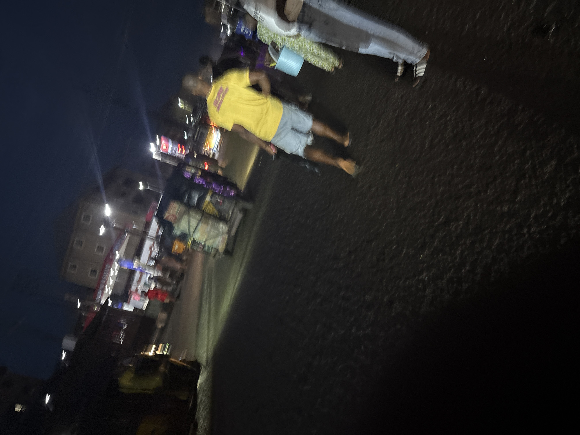 This is a nighttime street scene in Nkpor, Nigeria. A man in a yellow t-shirt and denim shorts walks past a blue bucket on the right. To the left, a bright yellow vehicle, possibly a rickshaw or small bus, is parked. In the background, a building with illuminated windows suggests a commercial area. Several other figures and vehicles are present, but they are blurred due to motion and low light conditions. The street surface is dark and appears wet, reflecting some of the light sources. The sky is dark blue, indicating late evening or night.