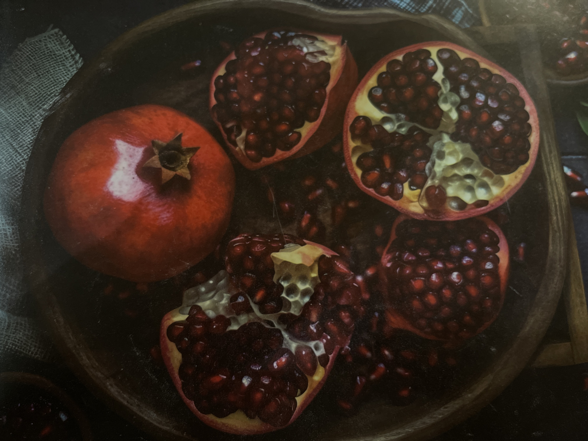 This media file is a photograph of pomegranates. The main subjects are several whole and cut pomegranates, arranged in a wooden bowl. Some of the pomegranates are whole and red, while others are cut open, revealing their numerous arils (seeds).
The scene is a still life, likely arranged for a photograph. The setting appears to be indoors, with a dark background and a textured burlap cloth on the left side of the frame. The lighting is dramatic, creating highlights on the smooth skin of the whole pomegranates and casting shadows, which adds depth to the composition.
There are no people or animals visible in the image. The activity taking place is implied by the presentation of the fruit – it is ready to be eaten or perhaps used as an ingredient. The time of day and weather are not discernible from the image. There are no visible text elements. The visual focus is entirely on the texture, color, and arrangement of the pomegranates. The composition is close-up, allowing the viewer to appreciate the details of the fruit, such as the glistening arils and the texture of the skin.