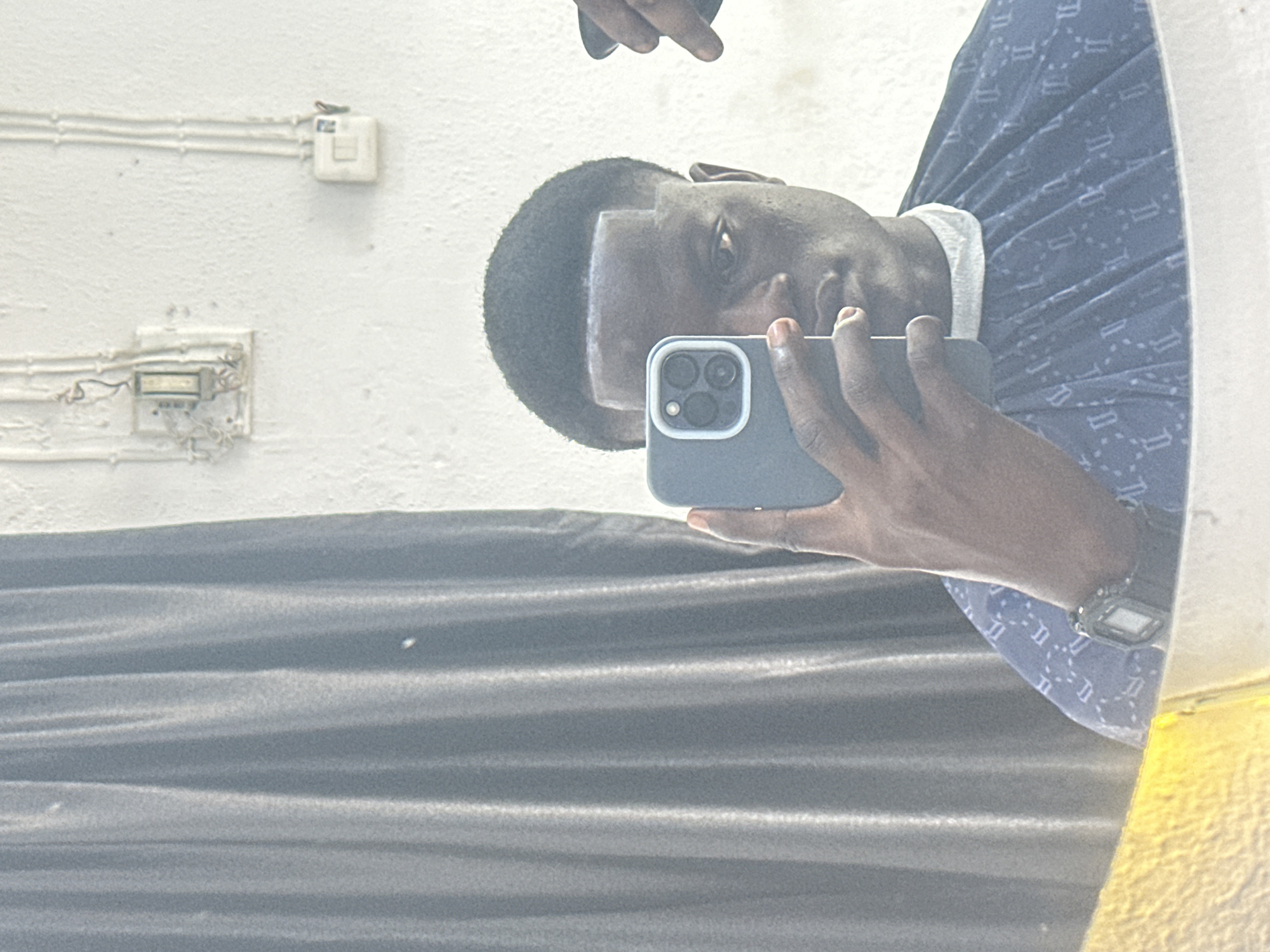 In the city of Badagry, Nigeria, a young man is captured in a mirror selfie, showcasing a fresh haircut. He holds a dark-colored smartphone in his right hand, angled to capture his reflection. The background reveals a textured white wall with exposed electrical conduits and a light switch, indicative of a utilitarian space, possibly a barber shop or a simple residence. To the left, a dark grey curtain hangs, adding a touch of texture and depth to the scene. The man is wearing a dark blue barber's cape with a white trim, printed with a subtle pattern. A black digital watch adorns his left wrist. The lighting is somewhat dim, casting subtle shadows and highlighting the contours of his face and hairstyle.
