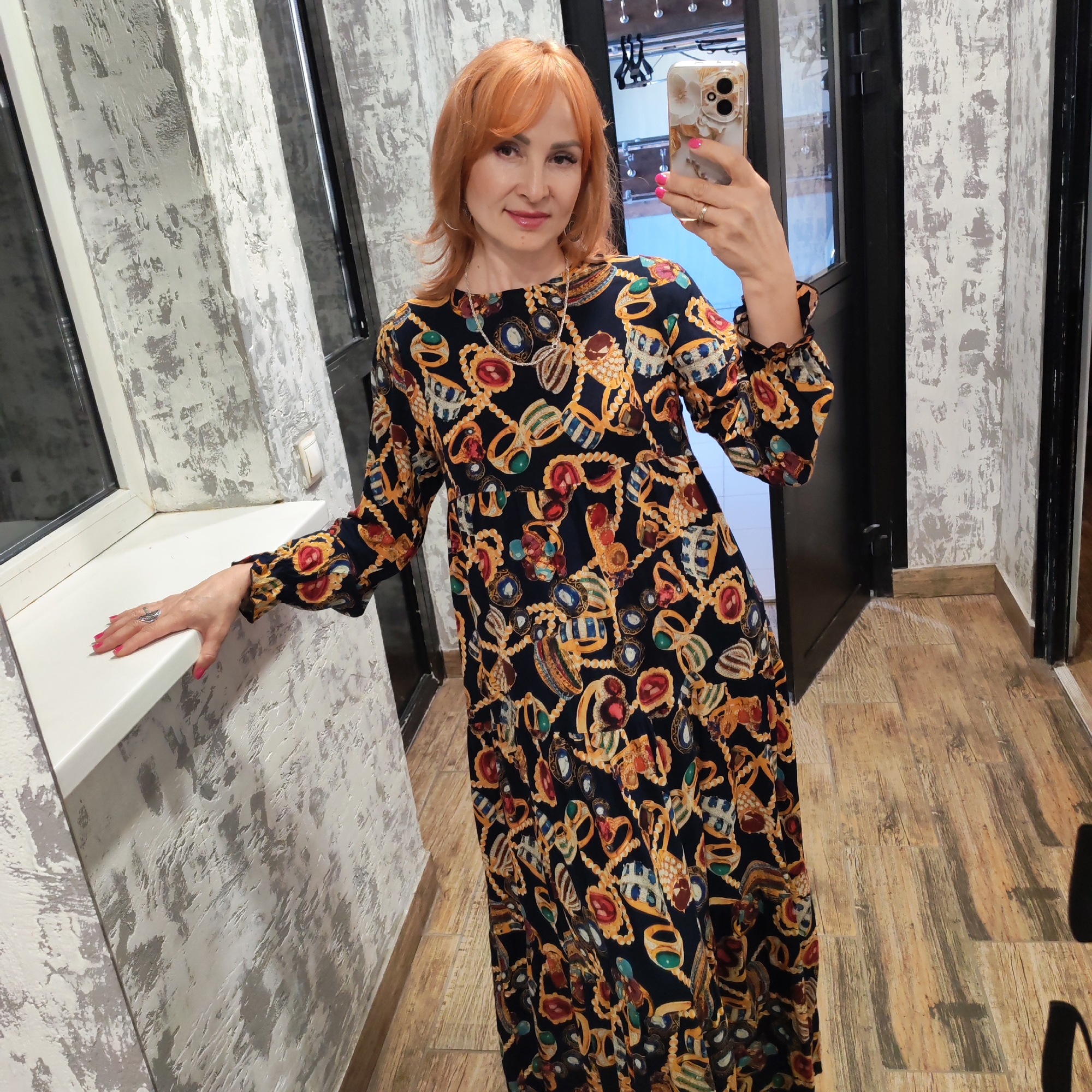 A middle-aged woman with reddish-orange hair is taking a selfie with her mobile phone in what appears to be a corridor. She is wearing a long, dark-colored dress with a jewelry-themed print and a silver necklace. She is leaning slightly against a white windowsill to her left. The wall behind her has a textured gray and white finish. The floor is made of light brown wood-like tiles. There is a doorway on the right with a dark frame, and what looks like a reflection in a mirror at the end of the corridor. Based on the available information, the city is Chaikovsky, Russia.