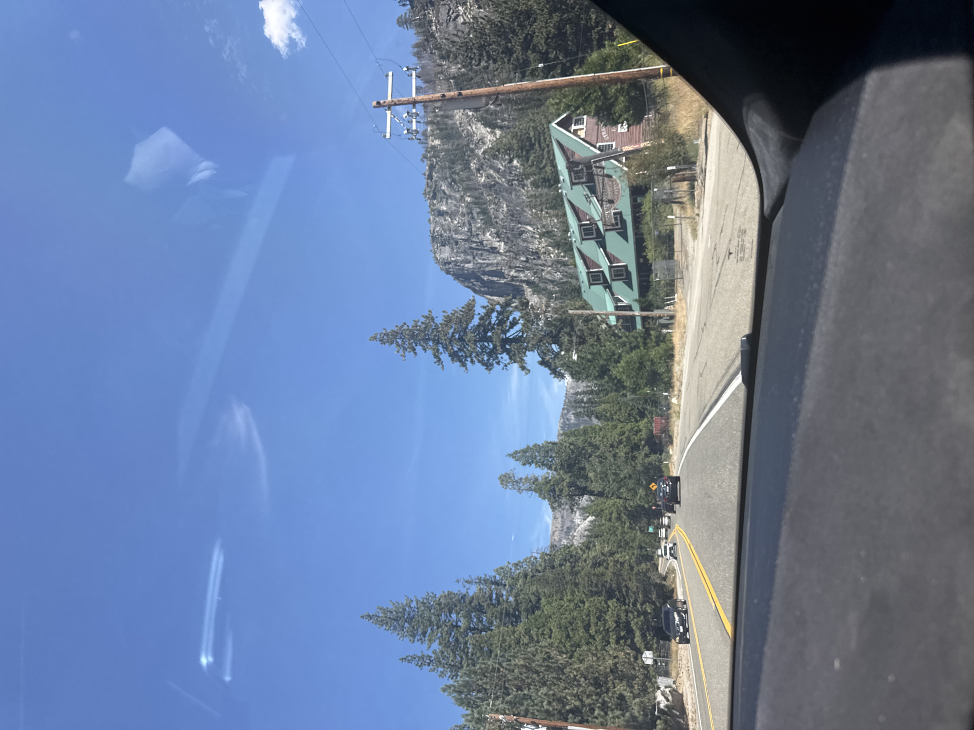 A highway curves through a mountainous, forested area under a clear blue sky. Tall, dark green pine trees line both sides of the road. On the right side of the highway, a large, rustic-style building with a green roof and brown walls is visible. The building has multiple dormer windows and appears to be a lodge or hotel. In front of the building, there is a dirt area with some fencing. Further up the hill behind the building, a steep, rocky mountain face is prominent. A wooden utility pole with power lines stands near the building. Several cars are driving on the highway, heading away from the camera. The image is taken from inside a vehicle, looking out through the windshield.