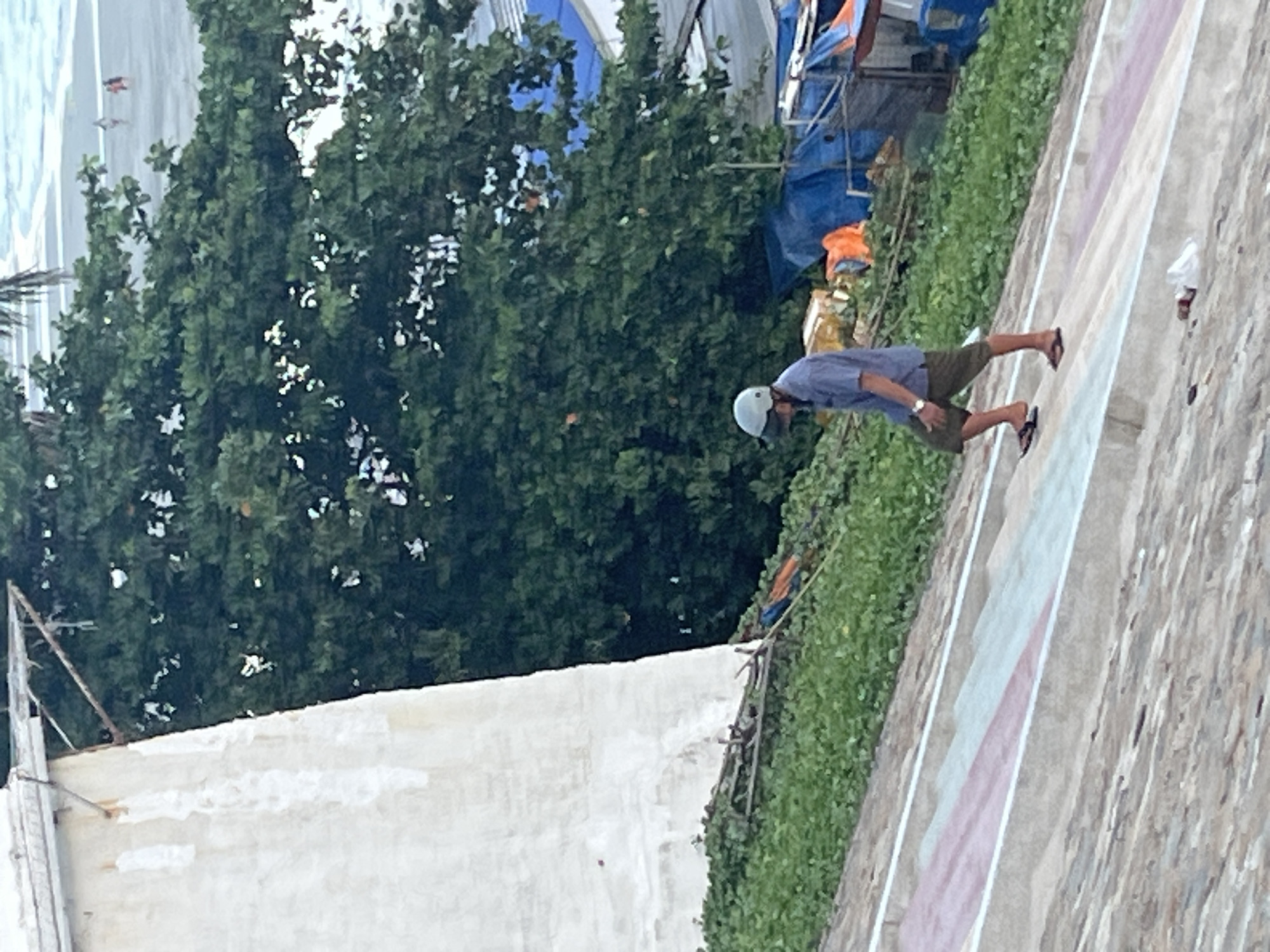 In Mũi Né, Vietnam, a man wearing a blue short-sleeved shirt, dark shorts, a helmet, and flip-flops walks up a concrete incline with painted stripes. He is walking away from the camera, towards a dense green tree line and the ocean. Behind the trees, some blue tarps and what appear to be makeshift structures are visible. The incline itself is made of rough concrete, with sections painted in pink, white, and light blue stripes. The lower portion of the incline is a rougher, more textured concrete. A small white object, possibly plastic, lies on the lower part of the incline.