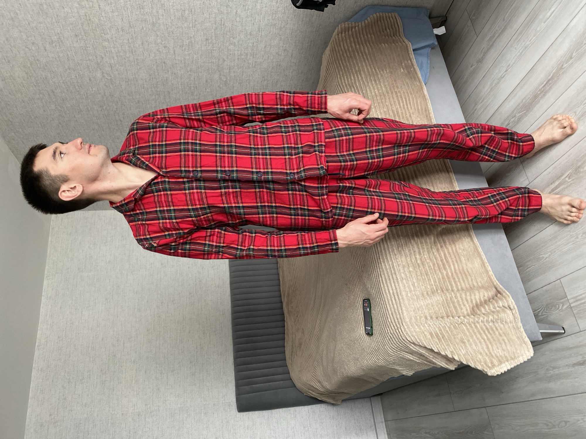 A man lies supine in a red plaid pajama set on a bed with a gray upholstered headboard and a light brown textured blanket. The bed is placed on a light gray wood-look floor, and a wall of similar color is visible behind the headboard. The location is Izhevsk, Russia.