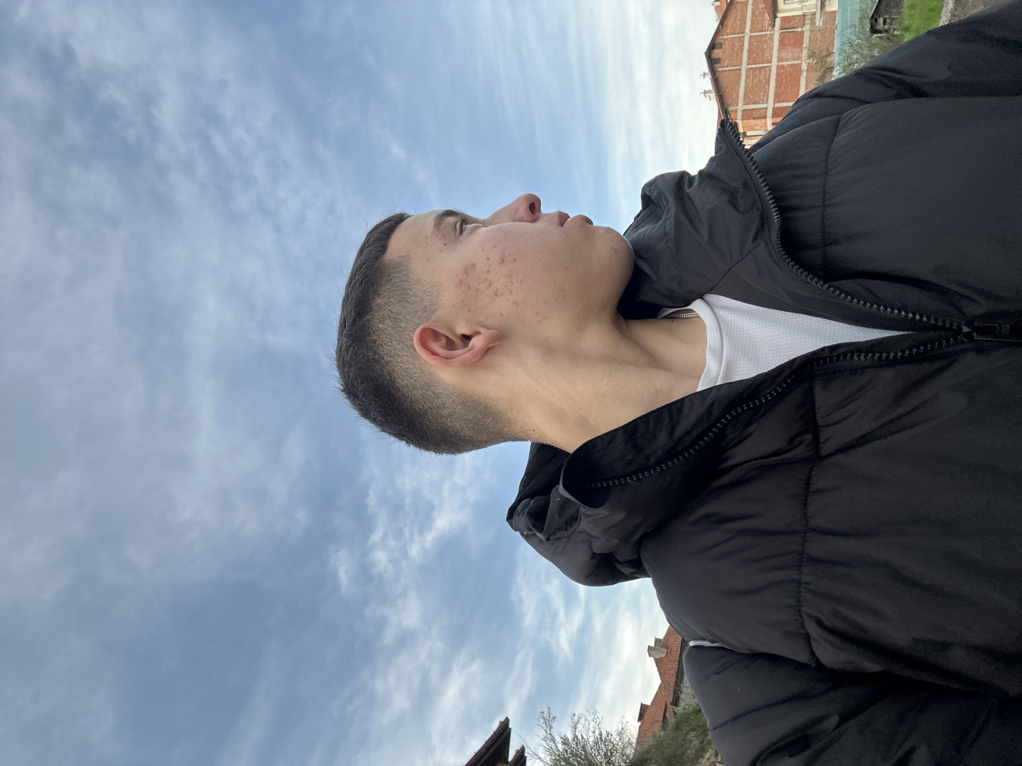 The image shows a young man wearing a black puffer jacket and a white t-shirt, standing outside in Zlatograd, Bulgaria. He is looking upwards towards the sky, which is cloudy with patches of blue. The buildings in the background appear to be made of brick and are typical of the architecture found in Zlatograd. The photo is taken from a low angle, emphasizing the sky and the man's upward gaze.