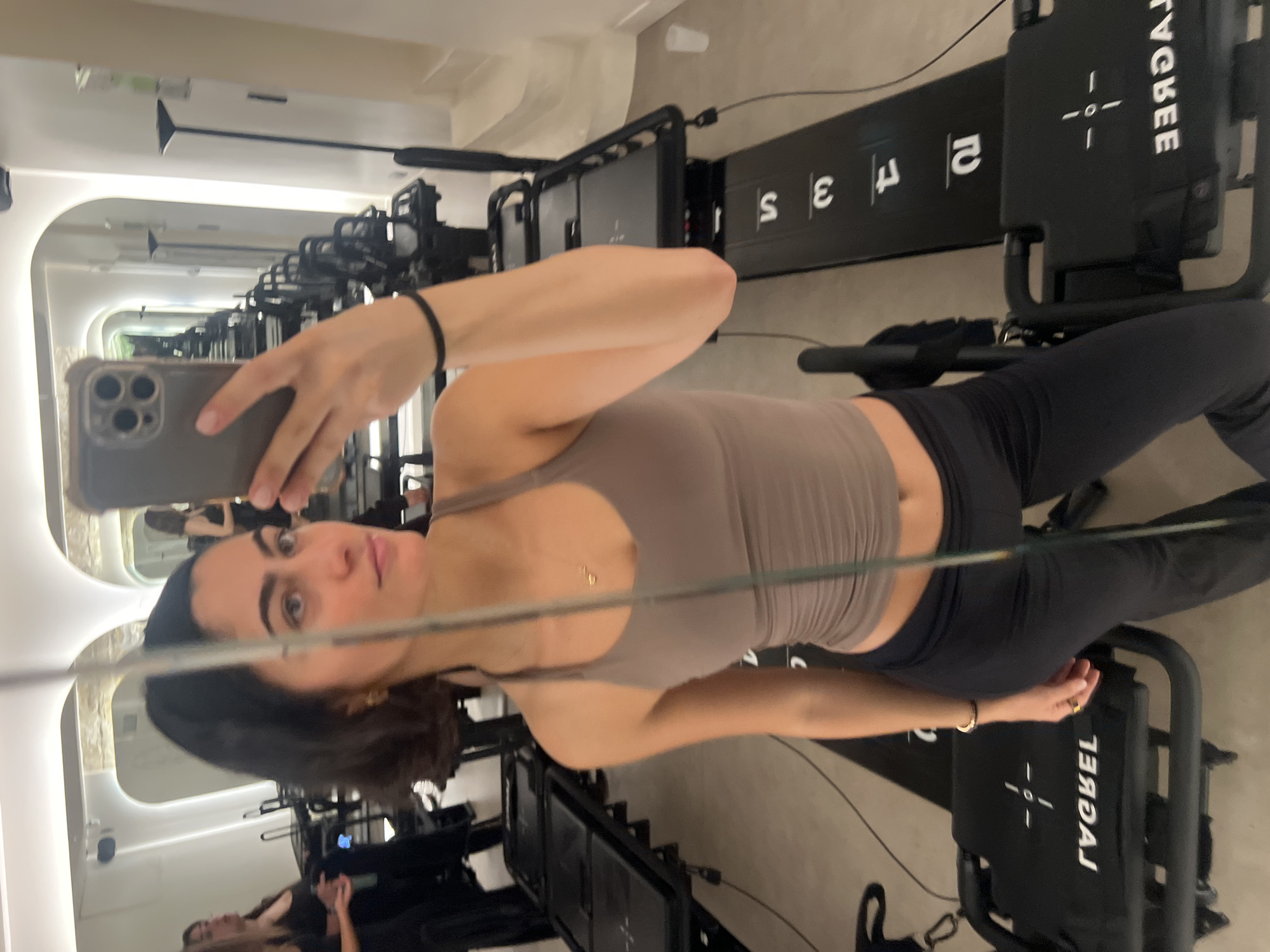 A woman is taking a selfie in a mirror, likely at a Lagree studio in Paris, France, given the presence of several Lagree machines visible in the background. The woman is wearing workout attire, a beige tank top and black leggings. She's holding a smartphone in her left hand, angled towards the mirror to capture her reflection. The reflection shows a line of Lagree machines extending into the background, creating a visual depth effect. The environment appears to be a clean, well-lit studio with a minimalist aesthetic. The mirror's reflection also captures glimpses of other individuals in the studio, possibly exercising or waiting for a class.