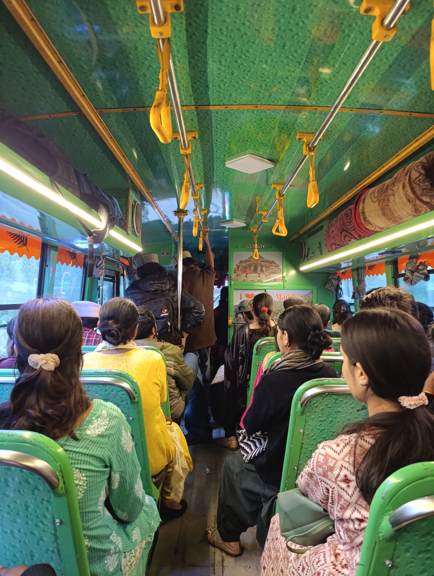 The image shows the interior of a passenger bus, likely in India given the provided context of Mansari. The bus features a distinctive vibrant green color scheme on its walls and textured seats, contrasted by bright yellow grab handles hanging from silver poles along the ceiling. Fluorescent strip lights illuminate the cabin, suggesting it might be dark outside or to enhance visibility.
Numerous people are seen inside the bus. Most passengers are seated, viewed from behind. In the foreground, a woman wears a light green and white patterned tunic, while another person wears a yellow top further back. To the right, another woman is dressed in a pink and white patterned outfit, carrying a green bag. Many female passengers have their hair tied back, some with scrunchies. Towards the front and middle of the bus, several men are standing and holding onto the overhead handles. One standing individual wears a black jacket and a backpack with the brand name "RAMBAT" visible on it.
The windows reveal a blurred, possibly rainy or misty, outdoor environment. Decorative or functional elements include rolled mats or rugs stored on an overhead rack on the right side of the bus. On the left side, posters are affixed to the bus wall. One poster prominently displays an image of a temple with the text "ANAND" above it. Below this, another poster reads "I ❤️ Manihar", indicating a potential local reference or destination. The scene captures a moment of public transport, with passengers on a journey or commute.