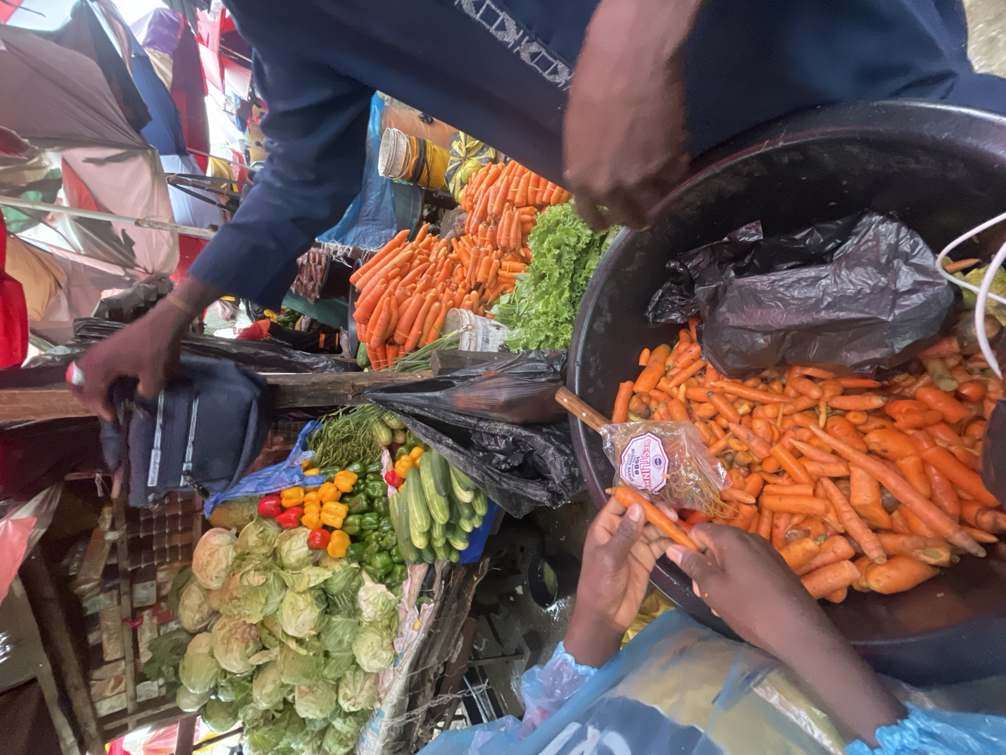 A high-angle shot depicts a bustling marketplace in Ado, Nigeria. The scene focuses on a vendor preparing carrots for sale. Carrots, both bundled and loose, dominate the view. A large black tub is filled with peeled carrots, while another pile of unpeeled carrots sits nearby. Additional produce, including cabbage heads, cucumbers, and peppers, is visible. Two individuals are present: one appears to be peeling carrots, while another is reaching for produce. The market is sheltered by tarps and makeshift roofing. A black bag sits in the large black tub.
