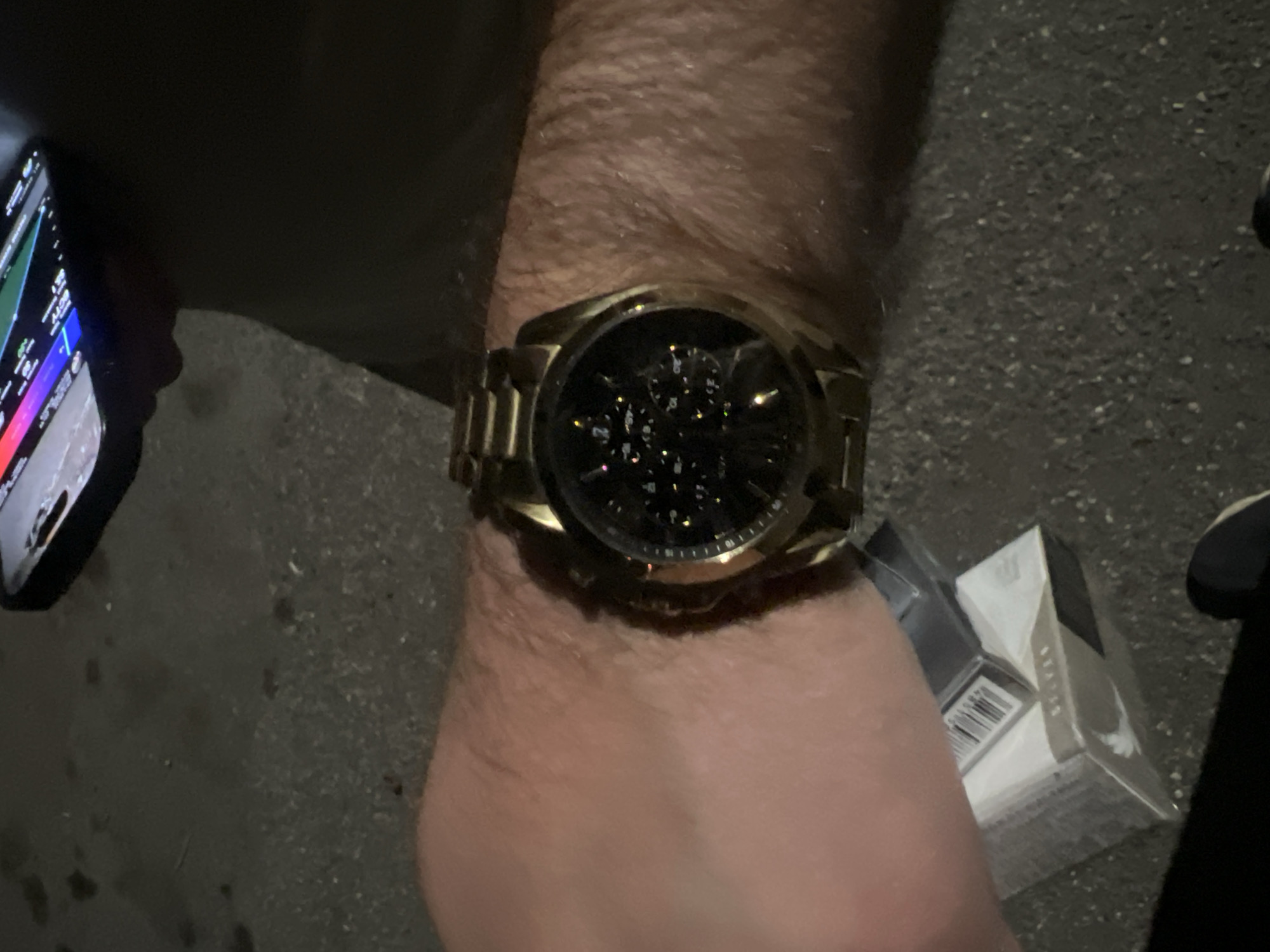 A close-up photograph taken outdoors at night. The subject is a person's left wrist, adorned with a gold-colored wristwatch with a dark face. To the left of the wrist is a smartphone with a display that is turned on, showing a colorful interface. To the right of the wrist is a partially visible object that is wrapped in plastic. The setting appears to be a paved surface, likely a sidewalk or street. The overall lighting is dim, contributing to the dark and somewhat grainy quality of the image. There is no explicit information available to indicate the city of Yerevan, Armenia.