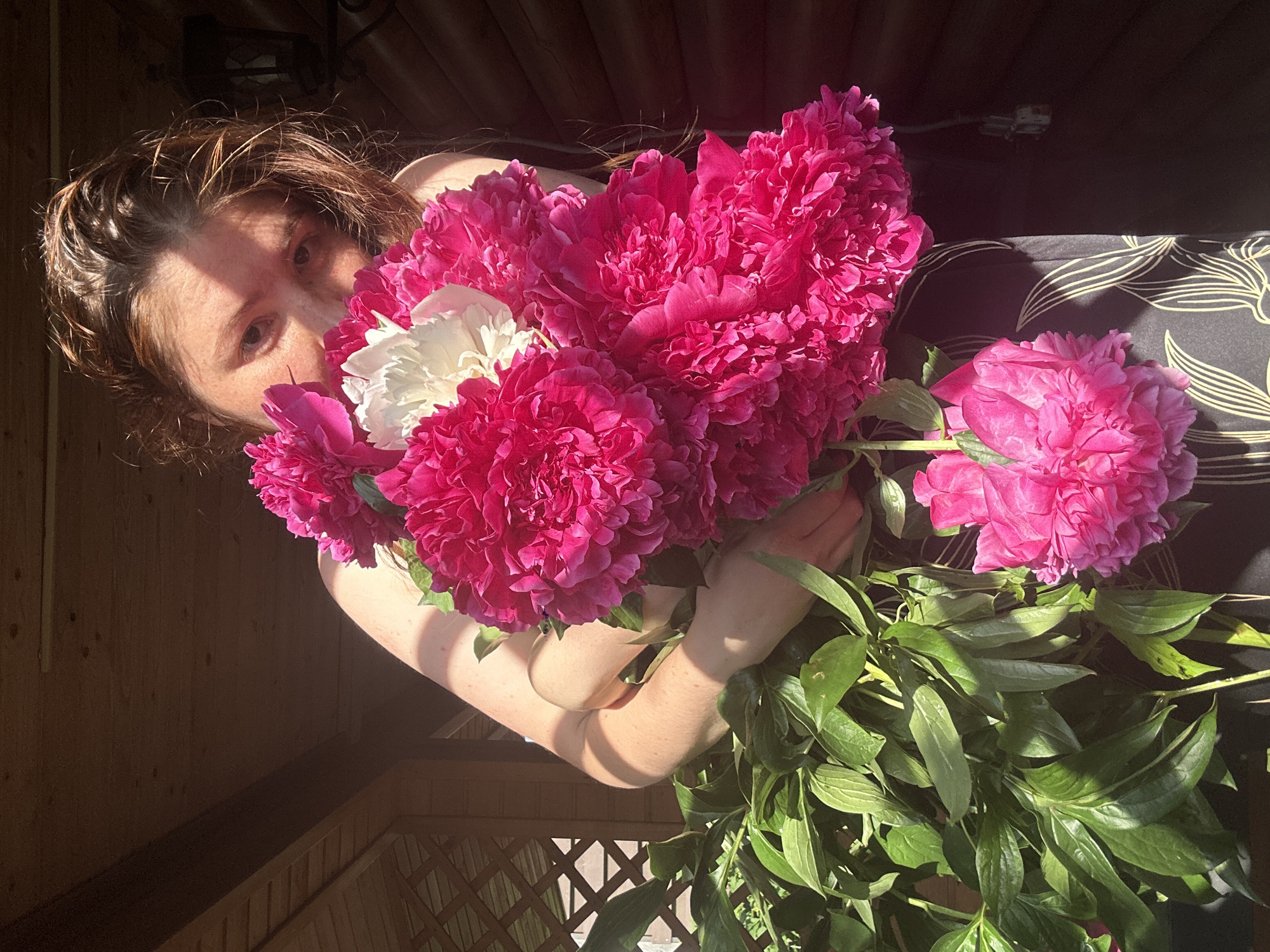 A person is holding a large bouquet of pink and white peonies, with their face partially obscured by the flowers. The person appears to be on a wooden porch or deck, with a wooden railing and a wooden wall visible in the background. The sunlight is casting shadows on the person's face and arms. The scene is consistent with a residential area in Nizhny Novgorod, Russia, during the peony blooming season.