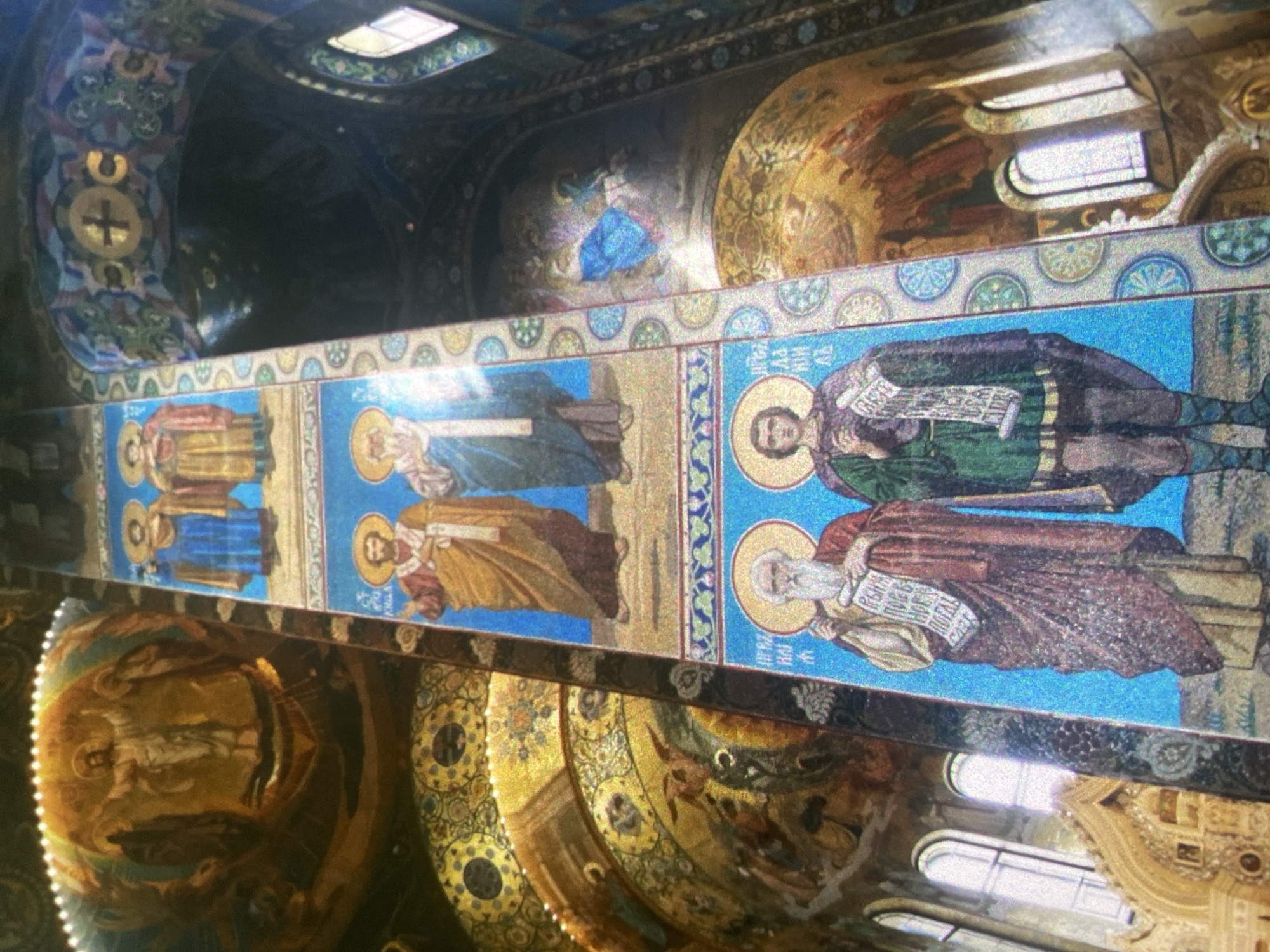 The image shows a section of the interior of the Church of the Savior on Spilled Blood in Saint Petersburg, Russia. The scene displays a mosaic frieze with several figures depicted in traditional Orthodox iconography. Each figure has a halo and is dressed in draped robes. They appear to be holding scrolls or books, possibly containing religious texts. The background of the frieze is a vibrant blue with a decorative border.
Above and below the frieze, there are other mosaics and decorative elements typical of Russian Orthodox church interiors. These include dome-shaped sections with depictions of religious figures in a golden color scheme, along with windows providing natural light. The image is taken from an oblique angle, offering a view of the intricate details and the overall opulence of the church's interior.