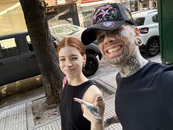 The image captures two individuals, a female and a male, posing for a selfie on an urban street in Buenos Aires, Argentina, during daytime. The female, on the left, has reddish-brown hair, a septum piercing, and wears a black tank top. She smiles with partially closed eyes and points her tattooed right index finger towards the camera. The male, on the right, has extensive facial and neck tattoos, large ear gauges, and wears a black baseball cap with various patches, along with a black t-shirt. He displays a wide smile, appearing to hold the camera. Behind them, a tree with rough bark stands on a checkered sidewalk. Parked vehicles are visible in the background, including a dark SUV to the left and a white SUV to the right, alongside buildings with windows and a metal rolling shutter. A green metal gate is partially visible on the far right.
