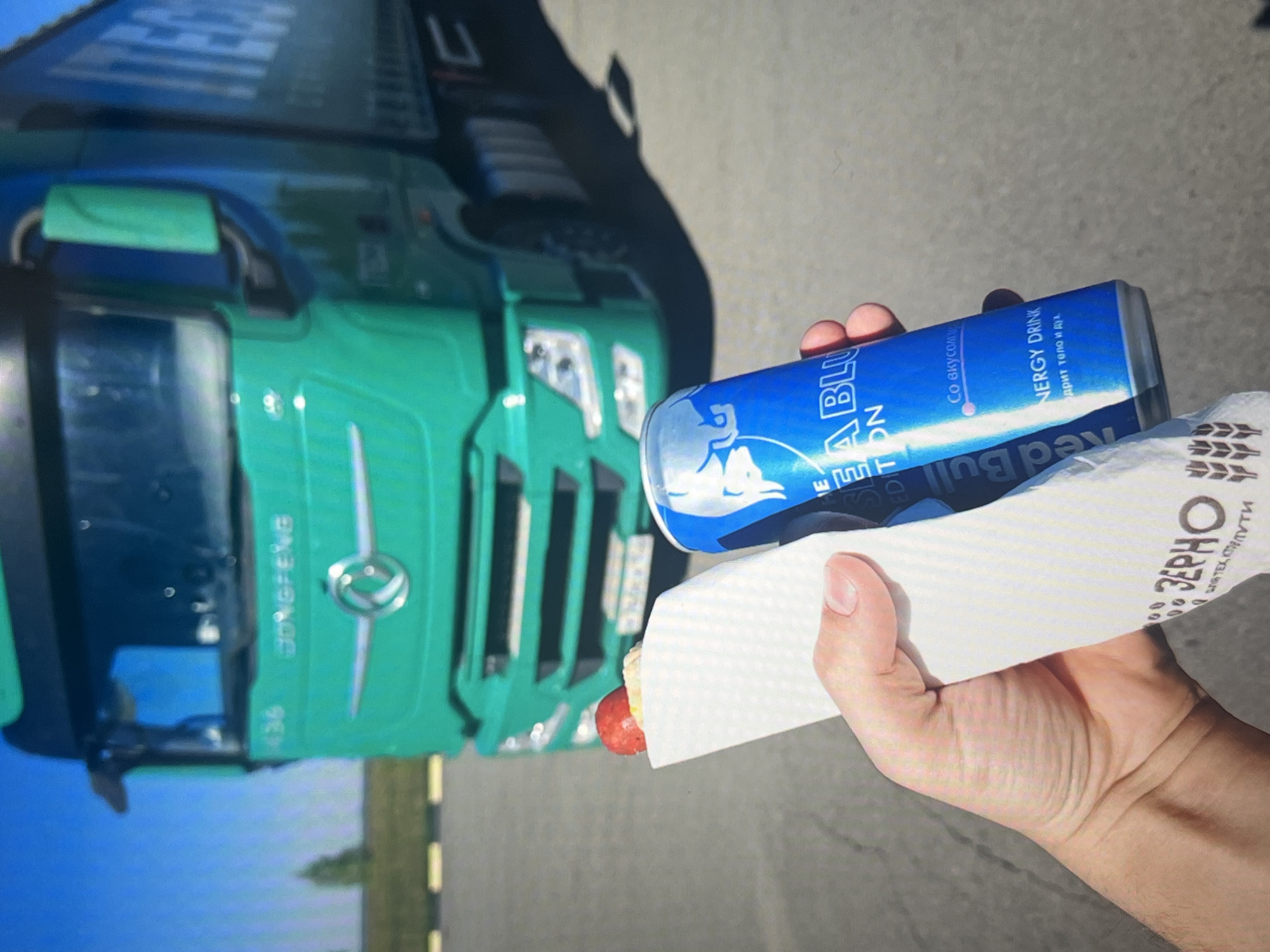 A person holds a hot dog and a can of Red Bull Blue Edition in front of a green Dongfeng truck parked on an asphalt surface. The truck is the primary subject in the background, with its grille and logo clearly visible. The hot dog is wrapped in white paper, and the Red Bull can is blue with the brand's bull logo. The setting appears to be an outdoor parking lot, possibly at a truck stop or rest area, with a clear blue sky overhead. The ground is paved with asphalt, and a few trees are visible in the distance. The image is taken from a slightly low angle, looking up at the truck and the person's hands holding the food and drink. The overall impression is a casual moment of refreshment during a journey. The location is identified as the city of Ob, Russia.