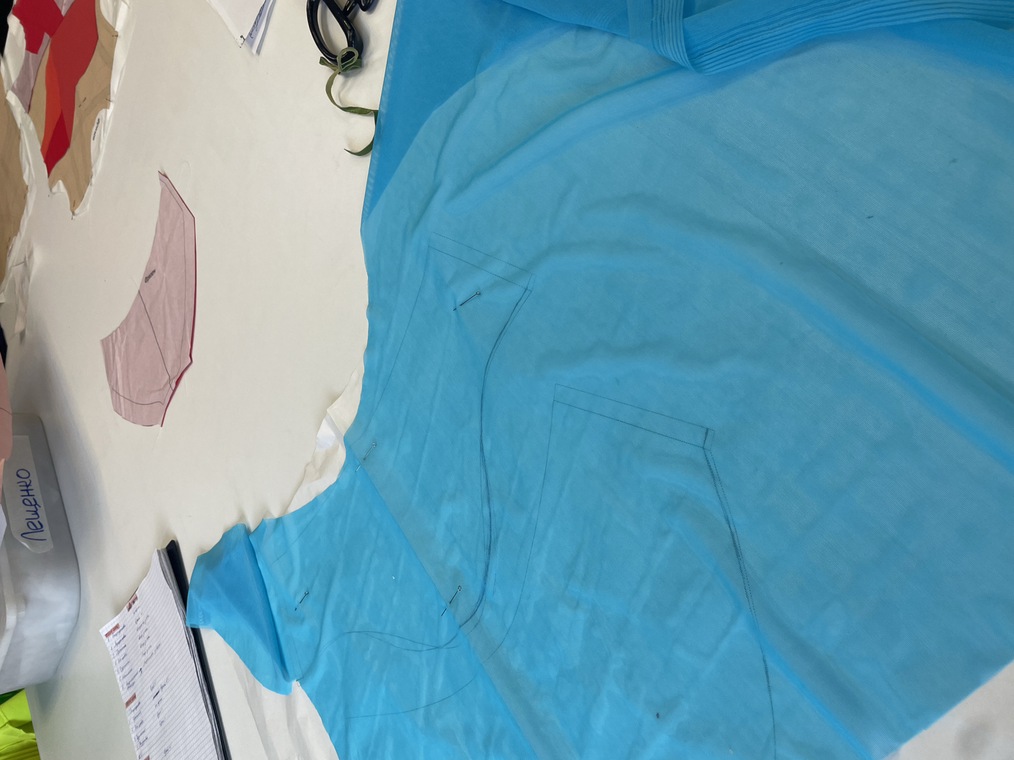 The image depicts a close-up view of a sewing table in what appears to be a tailoring or design studio. A large piece of translucent turquoise fabric is spread across the table, with some sections pinned. A pattern, possibly for a garment, is visible on the fabric. To the left, a notebook or ledger with lists and columns is present. In the background, a white plastic container with writing that appears to be Cyrillic script ("Лещенко") is visible, along with other fabric patterns in various colors. Scissors and a green ribbon are also on the table. The overall scene suggests a workspace where clothing is being designed or constructed. The request for the location in Sinyavino, Russia, cannot be fulfilled as the image itself provides no geographical information, and no external context or metadata suggests this location.