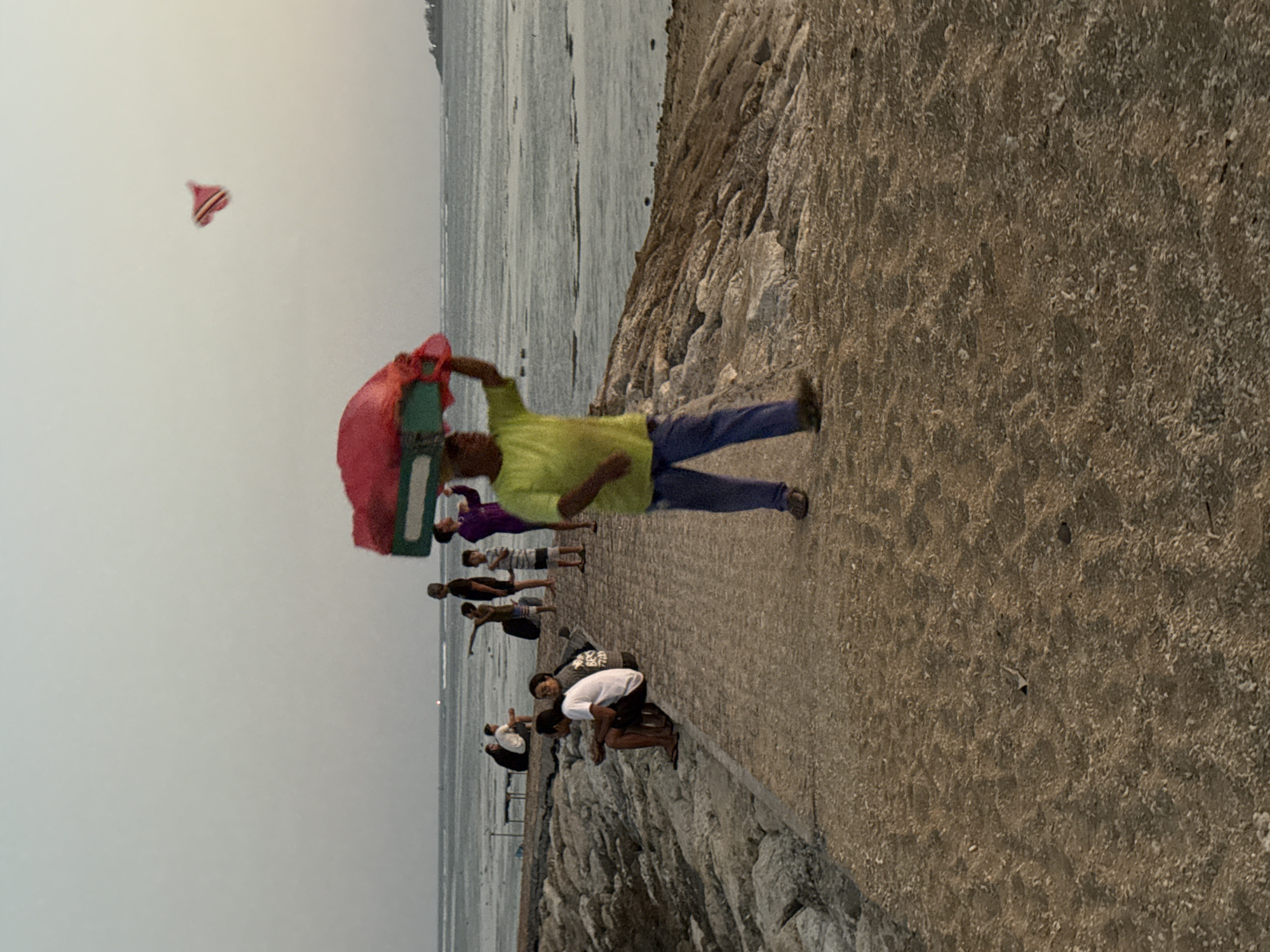 A person in a yellow shirt and blue pants carries a red and green object on their head while walking on a path beside a rocky shoreline. Several other people are visible in the background, some sitting and others standing. A kite is flying in the sky. The scene appears to be a public area near the water, possibly a beach or pier, in Denpasar, Indonesia. The overall lighting suggests it is either early morning or late afternoon.