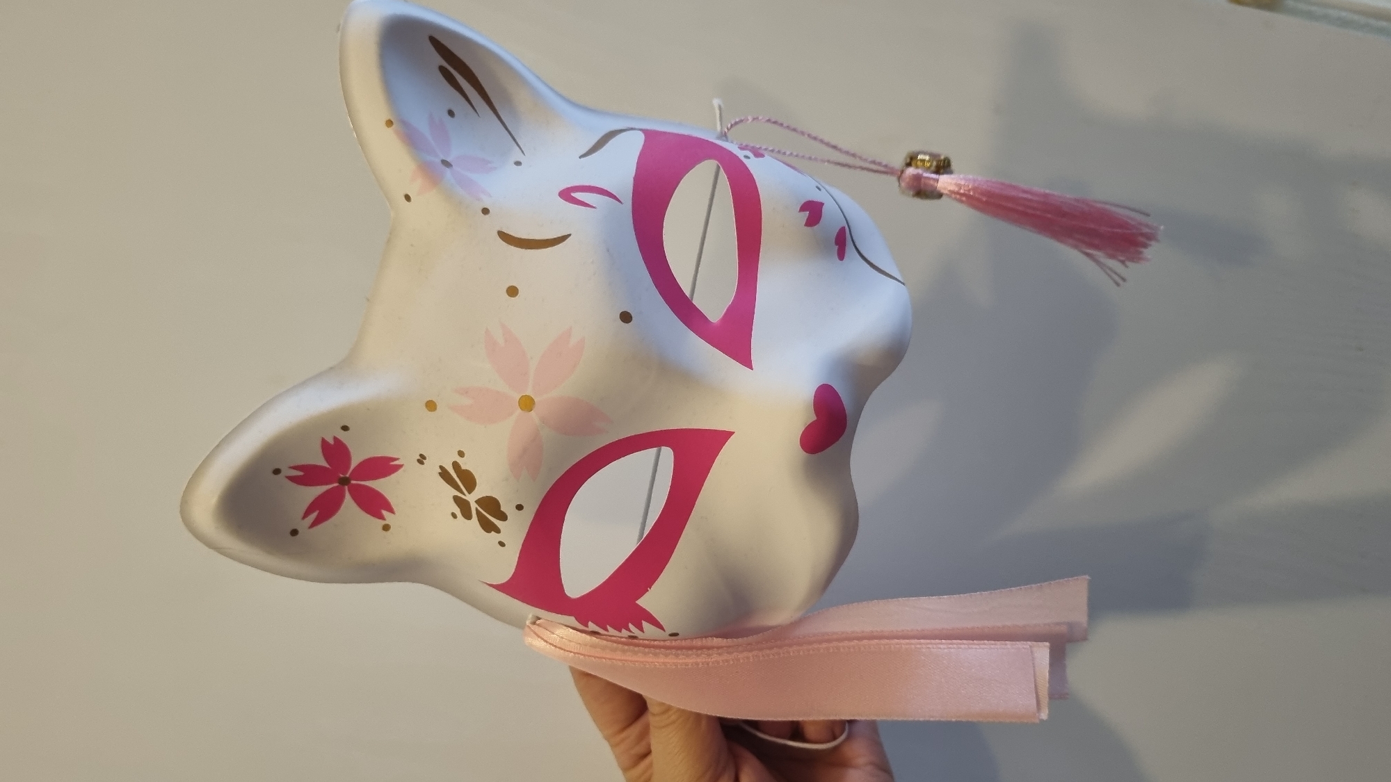 A white, fox-shaped mask with pink and gold cherry blossom designs is held in a person's left hand against a plain, light-colored wall. The mask has large pink almond-shaped eye cutouts, a small pink heart-shaped nose, and pink markings on the cheeks. A pink ribbon is attached to the left side of the mask, and a pink tassel with a gold bead hangs from the right side. The background is a neutral wall, casting a soft shadow from the mask. The image appears to be taken indoors, and there is no indication of its location in Moscow, Russia.