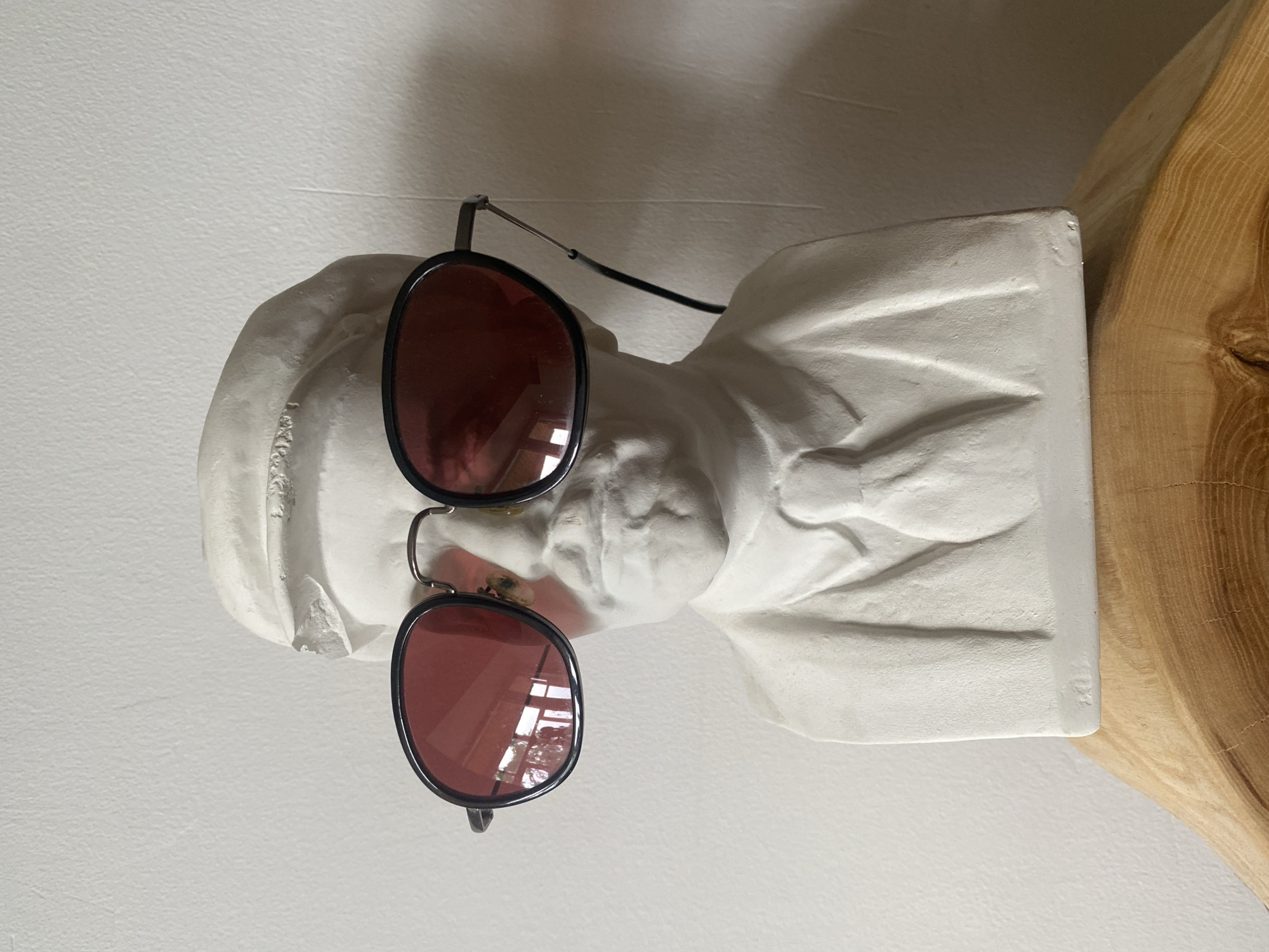A white plaster bust of a person with a shirt and high collar is photographed from a slightly high angle. The bust is wearing round, reddish-brown sunglasses. The glasses have black frames and silver earpieces. To the right of the bust, a section of light-colored wood is visible. The background is a plain, light-colored wall. The location is Rusanovo, Russia.