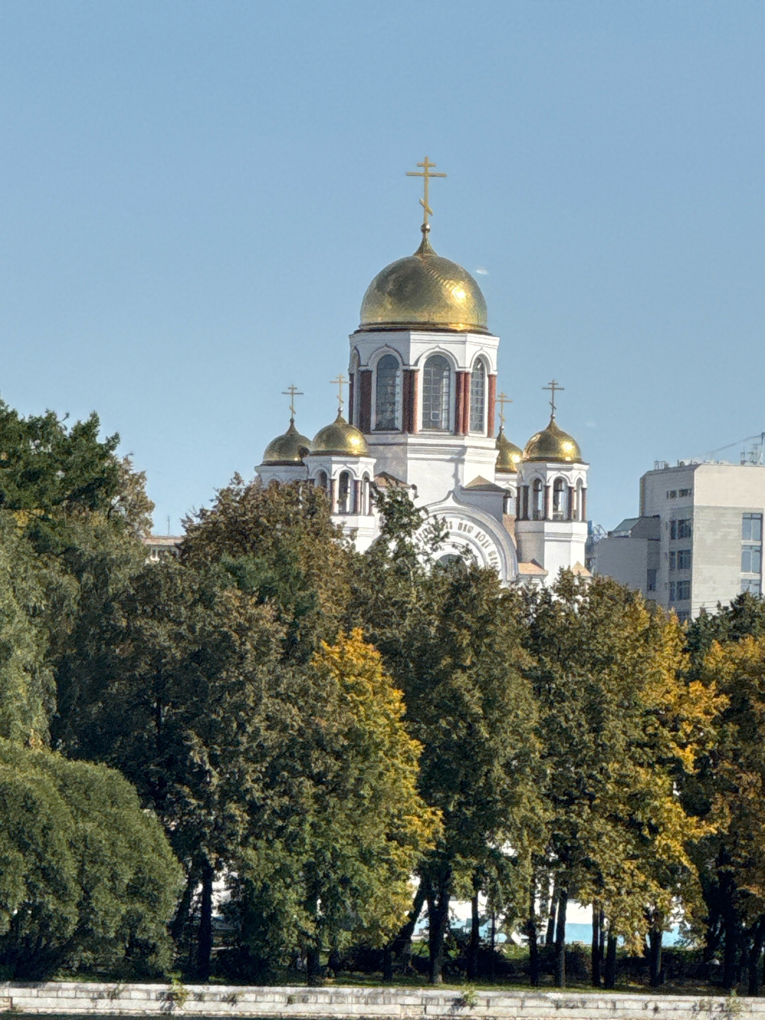 A golden-domed church, the Church on Blood in Yekaterinburg, Russia, stands tall against a clear blue sky. The church features white walls with red trim and several smaller golden domes topped with crosses. The foreground is filled with lush green trees, some of which are beginning to show autumn colors of yellow and orange. In the background, a modern, light-colored building is visible. The bottom of the image shows a stone wall or embankment. The scene appears to be a daytime, outdoor view.