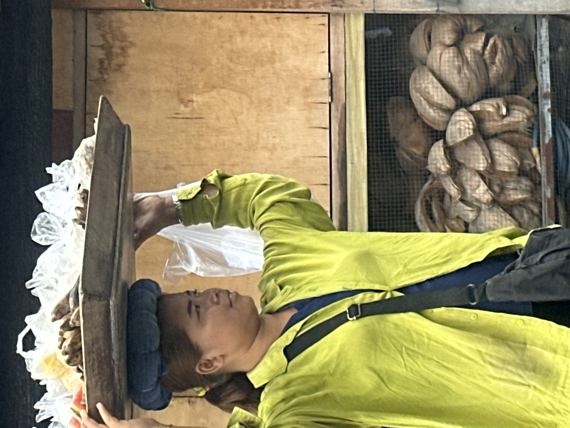 A woman in Denpasar, Indonesia, carries a tray of fruit and other items balanced on her head. She is wearing a bright lime-green, long-sleeved shirt over a dark blue undershirt and has a dark bag slung across her body. The tray on her head contains bananas, some watermelon slices, and other items wrapped in plastic bags. In the background, there is a wooden structure and a shelf with what appear to be coconuts or similar round objects visible through a mesh. The overall scene suggests a market or street vendor setting.