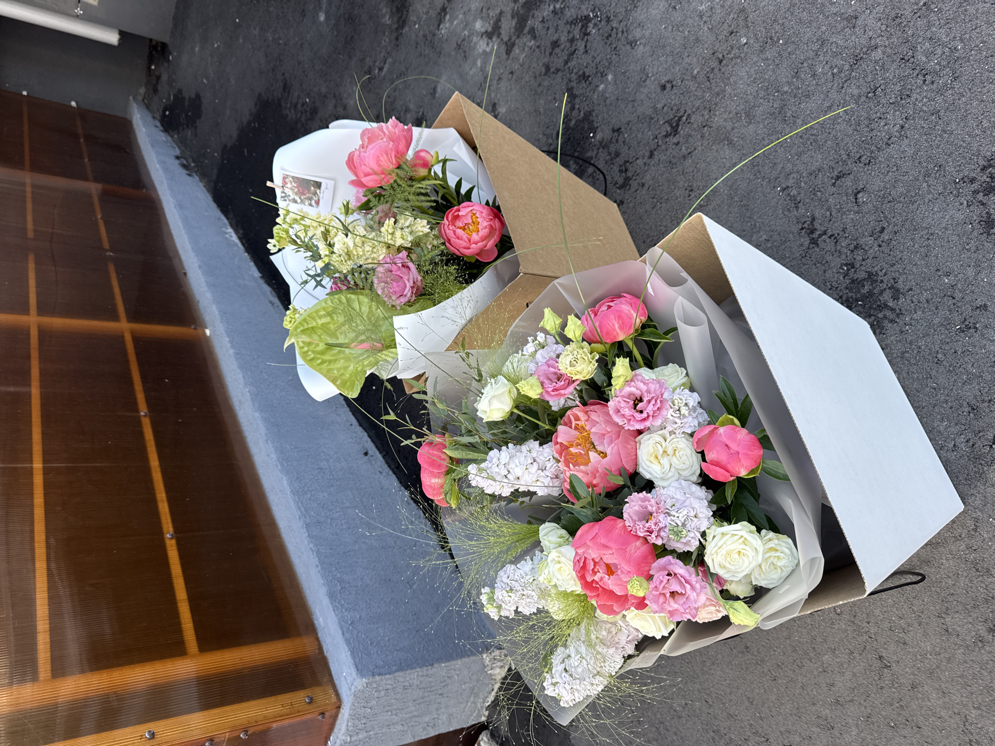Two flower bouquets are displayed in cardboard boxes on a grey asphalt surface. The bouquet in the foreground is in a white box and features pink peonies, white roses, and white hyacinths. The second bouquet, slightly behind and to the right, is in a brown cardboard box and includes pink peonies, white roses, green anthurium, and a small card attached with a clothespin. In the background, a grey concrete ledge runs along the base of a wall constructed with translucent orange panels held together by vertical wooden beams. The scene appears to be outdoors, possibly in a commercial or delivery staging area, but no specific Izhevsk, Russia location is identifiable from the visual evidence. No people or actions are depicted.