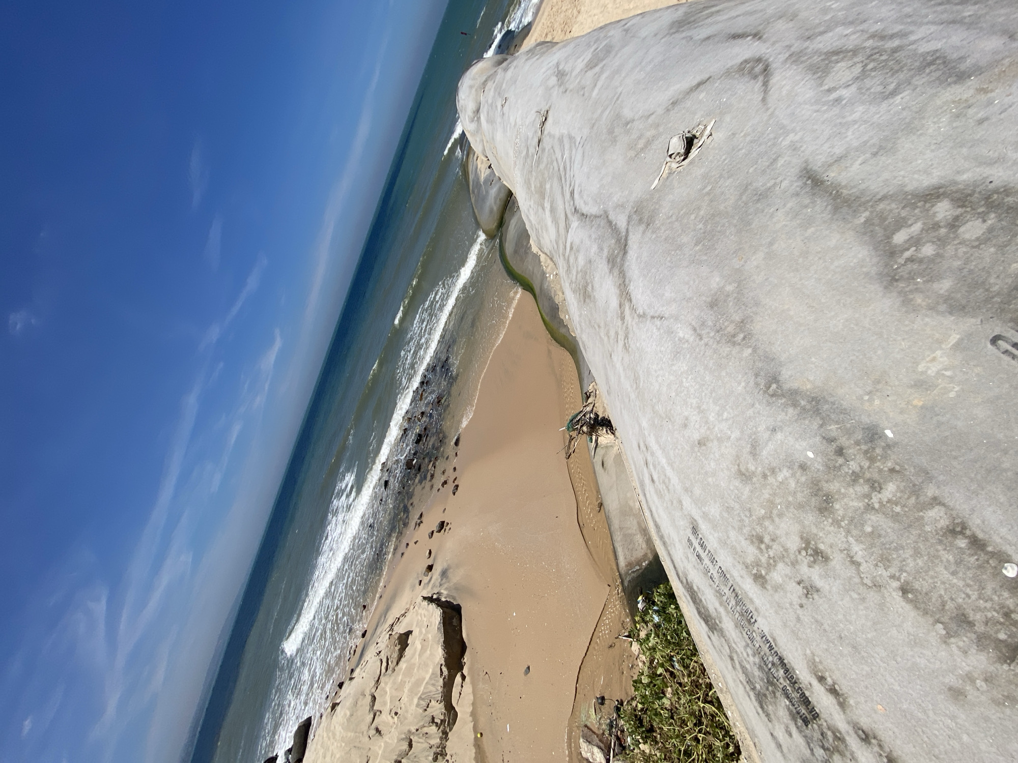 The image depicts a scene on a beach in Phu Thuy Ward, Vietnam. A large, weathered concrete pipe dominates the foreground, angled diagonally across the frame from the bottom right to the top left. The pipe's surface is textured and shows signs of age and exposure to the elements, with a faint inscription visible on its surface. To the left of the pipe, a patch of green vegetation grows, interspersed with scattered debris.
Beyond the pipe, a sandy beach stretches out, meeting the turquoise waters of the ocean. Gentle waves break on the shore, creating a foamy white line. The beach itself appears somewhat eroded in places, with exposed sand dunes and scattered rocks. A small stream of discolored water, possibly carrying sediment or debris, flows from the land towards the ocean, creating a sinuous green-brown line in the sand. The sky above is a clear, vibrant blue with a few wispy white clouds. In the distance, the coastline is visible, and a person can be seen standing in the water near the shore on the right side of the frame. The overall impression is one of a coastal environment, potentially impacted by infrastructure and natural erosion.