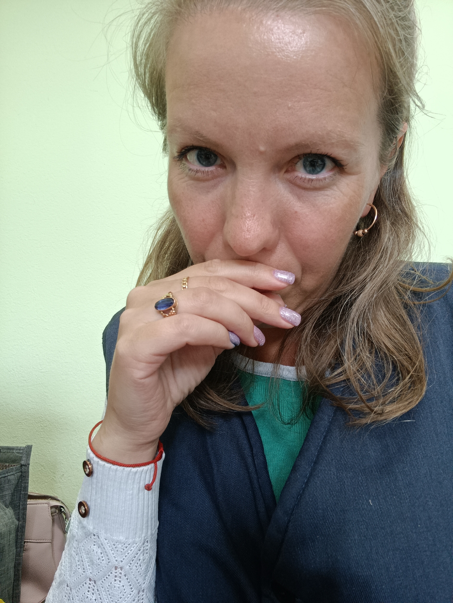 A woman is captured in a close-up, eye-level shot. Her eyes are blue. She has light blonde hair that is styled with slight waves. She is wearing a dark blue blazer over a green shirt. On her left hand, she has a gold ring with a blue stone and another ring with small details. Her fingernails are painted with a sparkly light purple polish. She also wears gold hoop earrings and a white sweater with decorative buttons and a red string bracelet. Her hand is partially covering her mouth and lower face. The background is a pale green wall. The image was taken in the city of Chaikovsky, Russia. The environment is indoors.