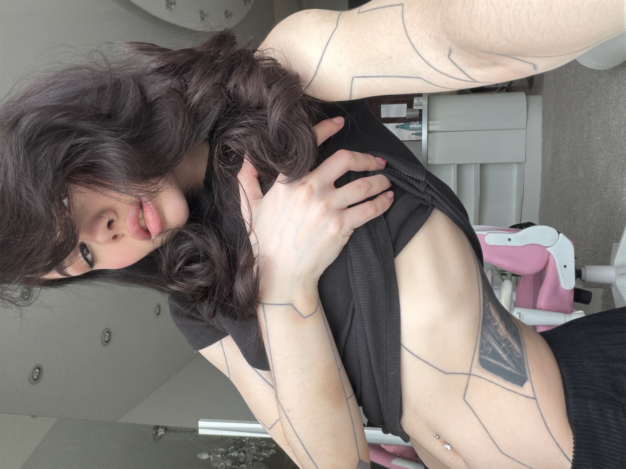 The image displays a person, likely female, with long, dark, wavy hair, captured in an indoor setting from a tilted perspective. The subject wears a black, short-sleeved top and dark bottoms. Their head is tilted, and they look towards the camera with a subtle open-mouthed expression, while voluminous hair partially covers one side of their face.
The person exhibits extensive body art, including geometric line tattoos covering their arms and extending onto the torso. A more elaborate, darker tattoo, possibly abstract or mechanical in design, is visible on the right side of the rib cage. A navel piercing with a clear gem is present on the abdomen. Traces of pink nail polish are visible on some fingernails.
The background suggests a bright, modern interior, likely a room in a home. Visible elements include a light-colored ceiling with multiple recessed lights, a chandelier, and various pieces of furniture. These include a white storage unit or shelving, and a prominent pink and white chair or high chair. The flooring appears to be a light-colored carpet or rug. The bright illumination indicates it is daytime. There is no visible text in the image. The scene conveys a stylized pose within a well-lit home environment, possibly in Khimki, Russia, as per context.